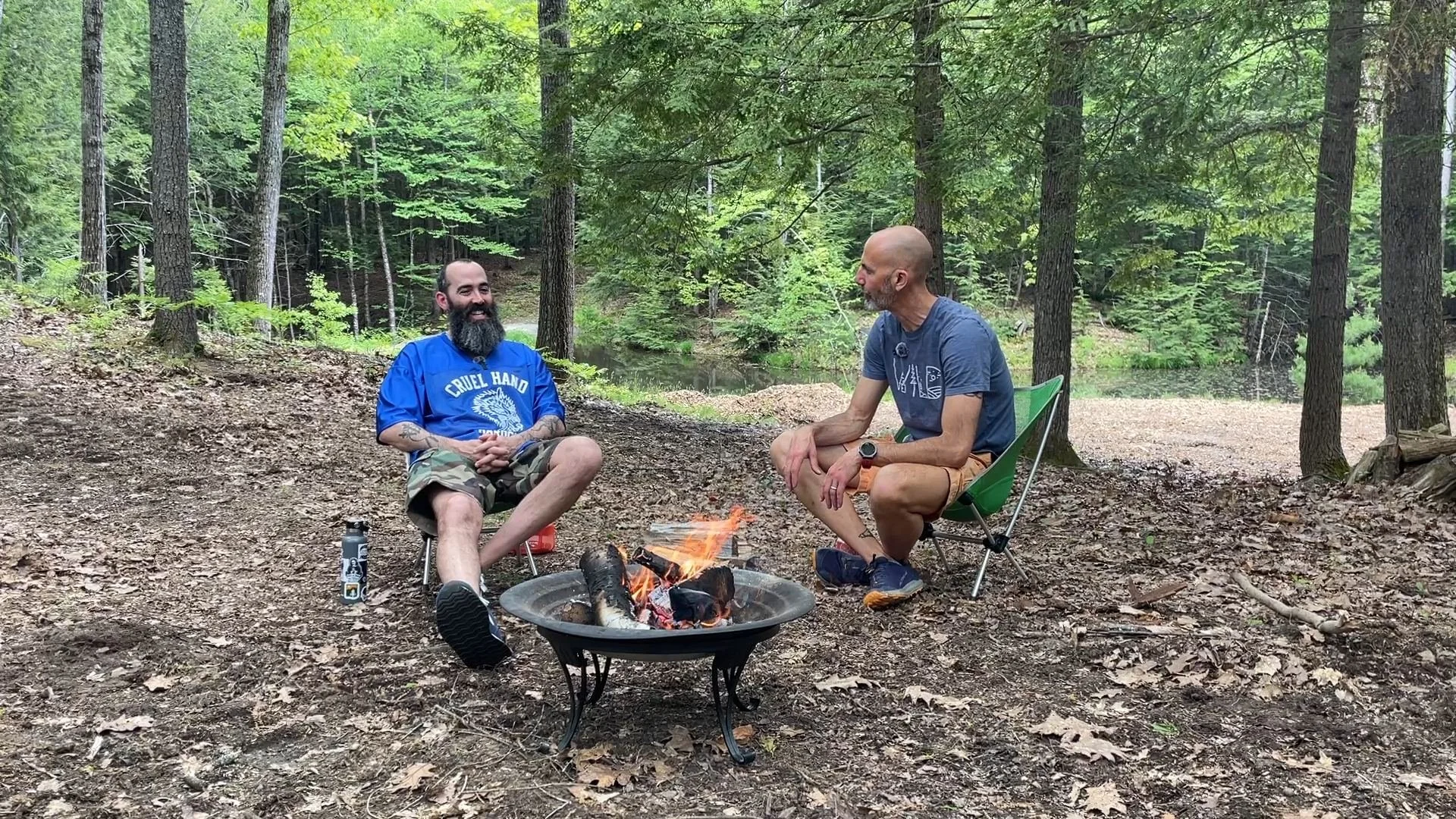 Two men sitting around a campfire in a forest, chatting, with trees and a body of water in the background.
