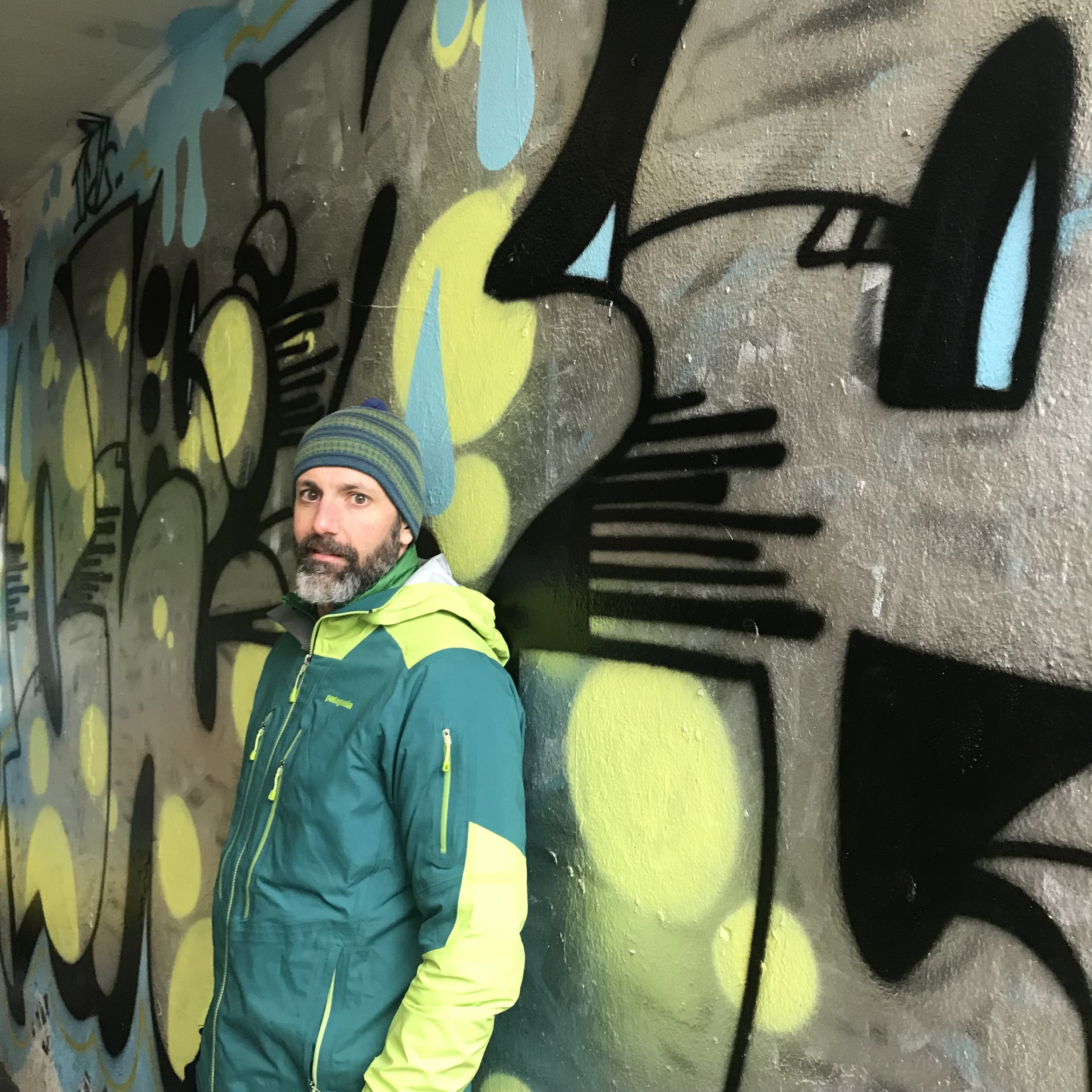 A man with a beard wearing a green and blue jacket and a striped beanie hat stands in front of a graffiti wall featuring abstract shapes and lines in yellow, blue, black, and gray.
