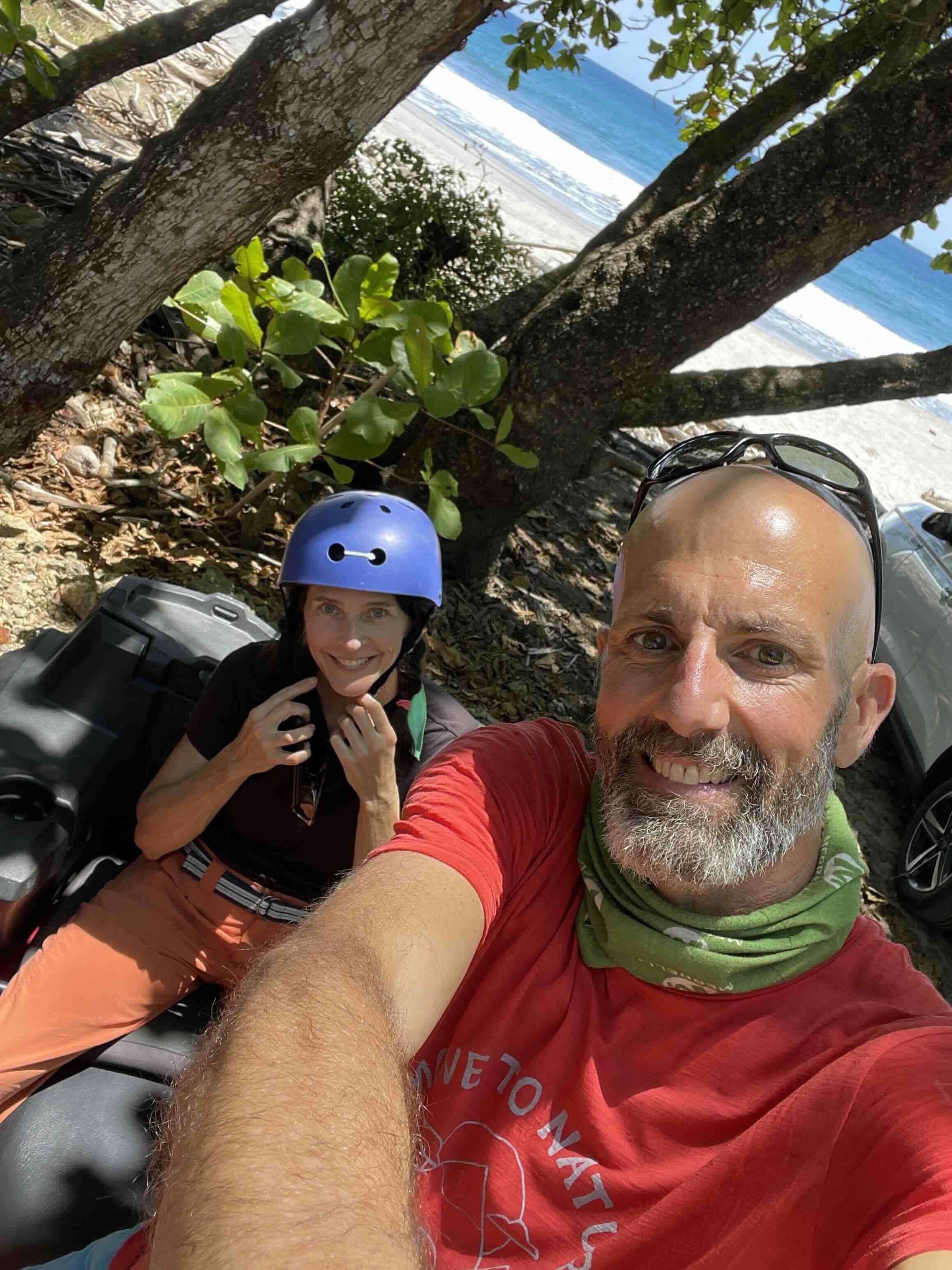 A man with a beard and sunglasses taking a selfie in front of a woman wearing a cap and sunglasses, with a beach and ocean in the background.