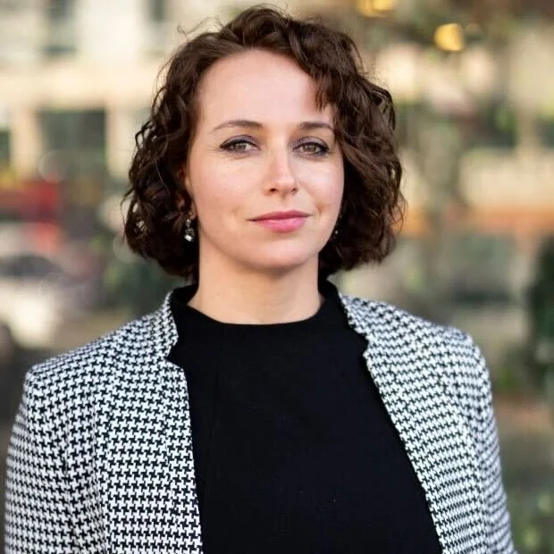 Woman with shoulder length hair is talking and looking away from the camera. She is wearing a patterned red, black and blue top.