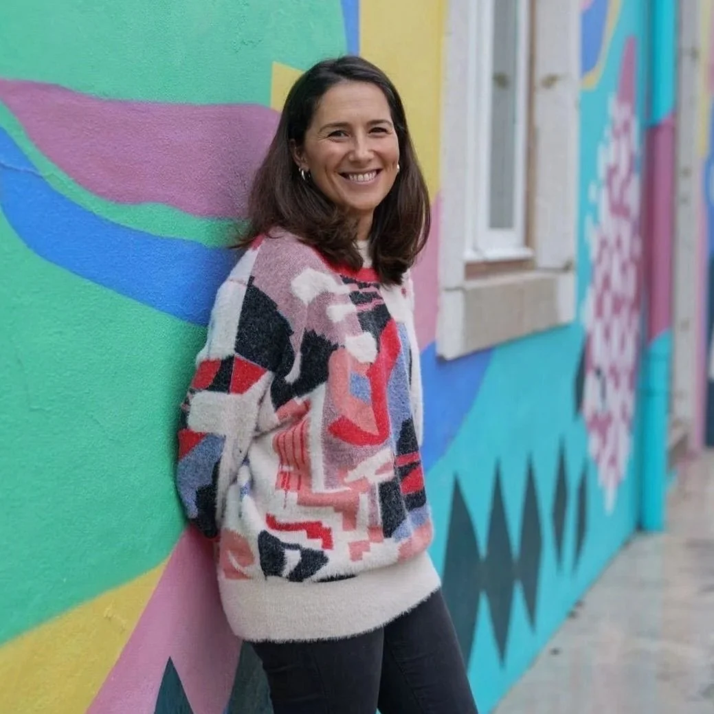 Smiling young woman with a colourful printed jumper, dark hair and light skin, outdoors leaning on a graffiti wall.
