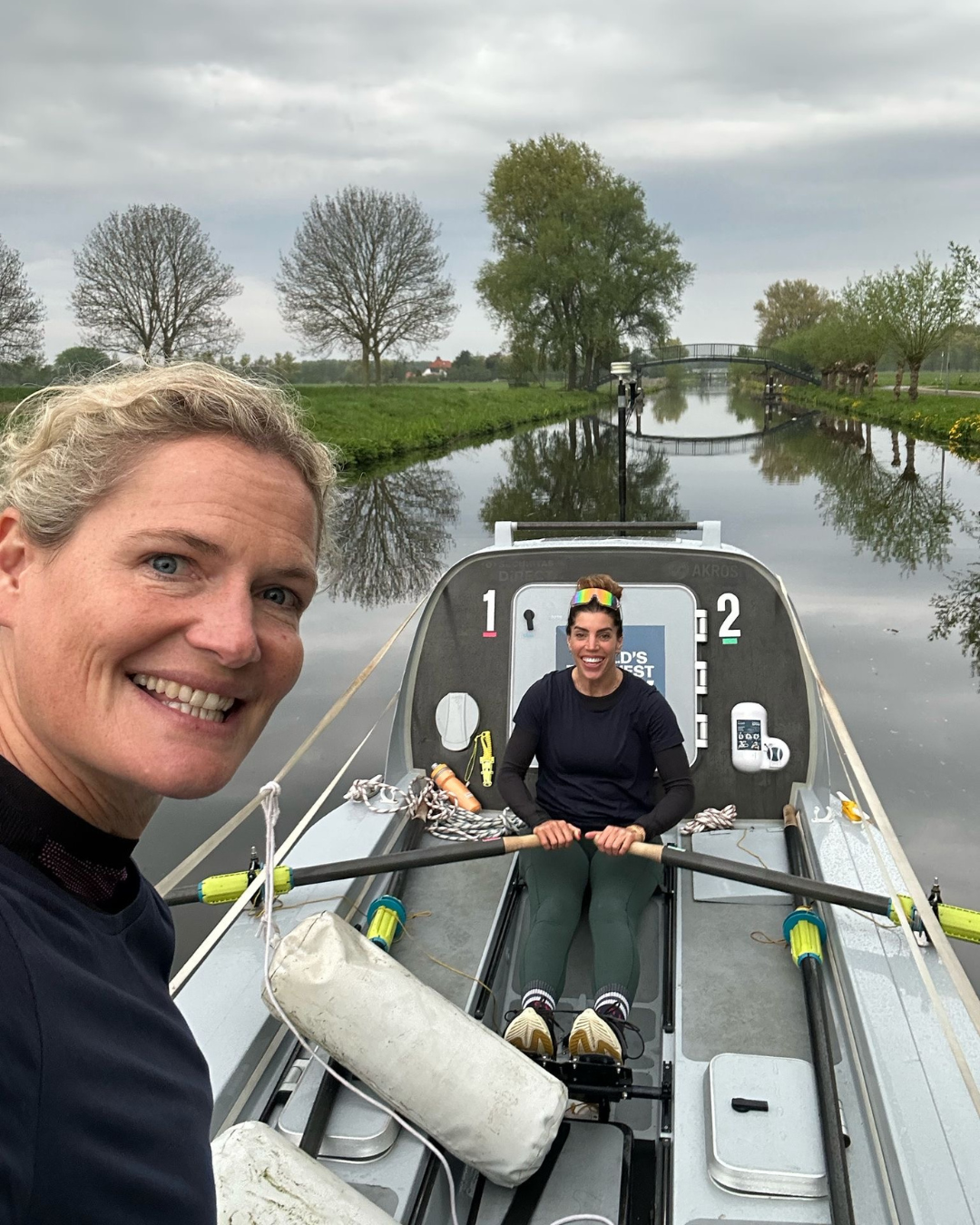 Two women are smiling to camera, one of them is rowing a small boat in the background while the other is standing taking a selfie image. You can see green fields and trees in the background