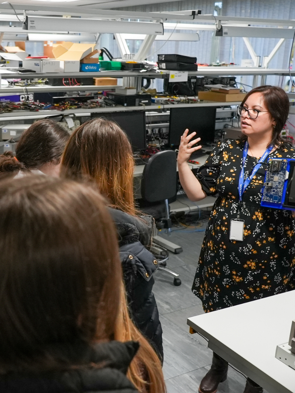 A woman giving a presentation or lecture to a group of people in a technical or engineering workspace, with computer monitors, electronic components, and equipment in the background.