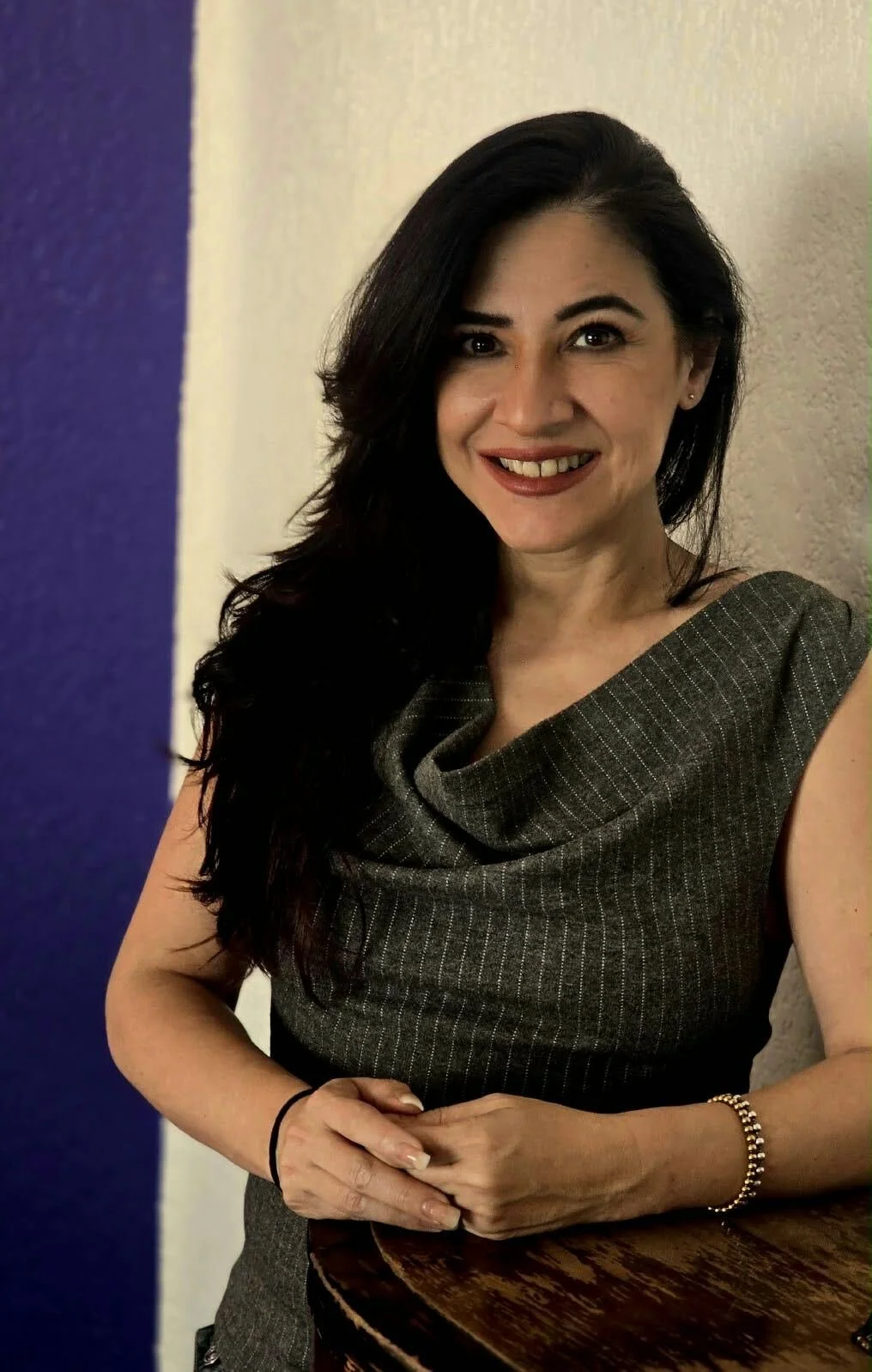 Woman is smiling to camera with long black hair, wearing a grey top,standing against a white and blue background.