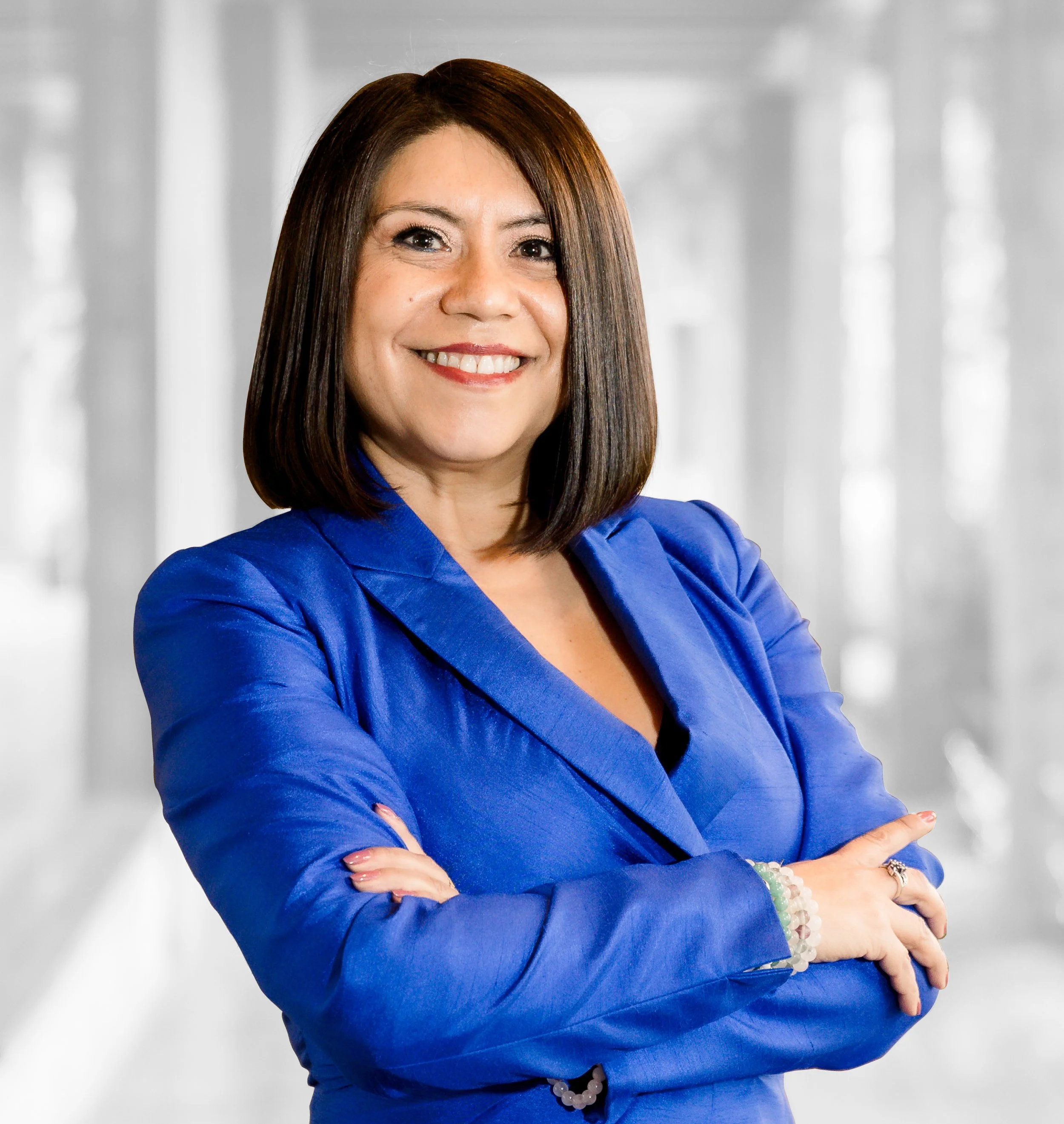Woman smile to camera, wearing a bright blue blazer and with her arms crossed. She has shoulder length brown hair.