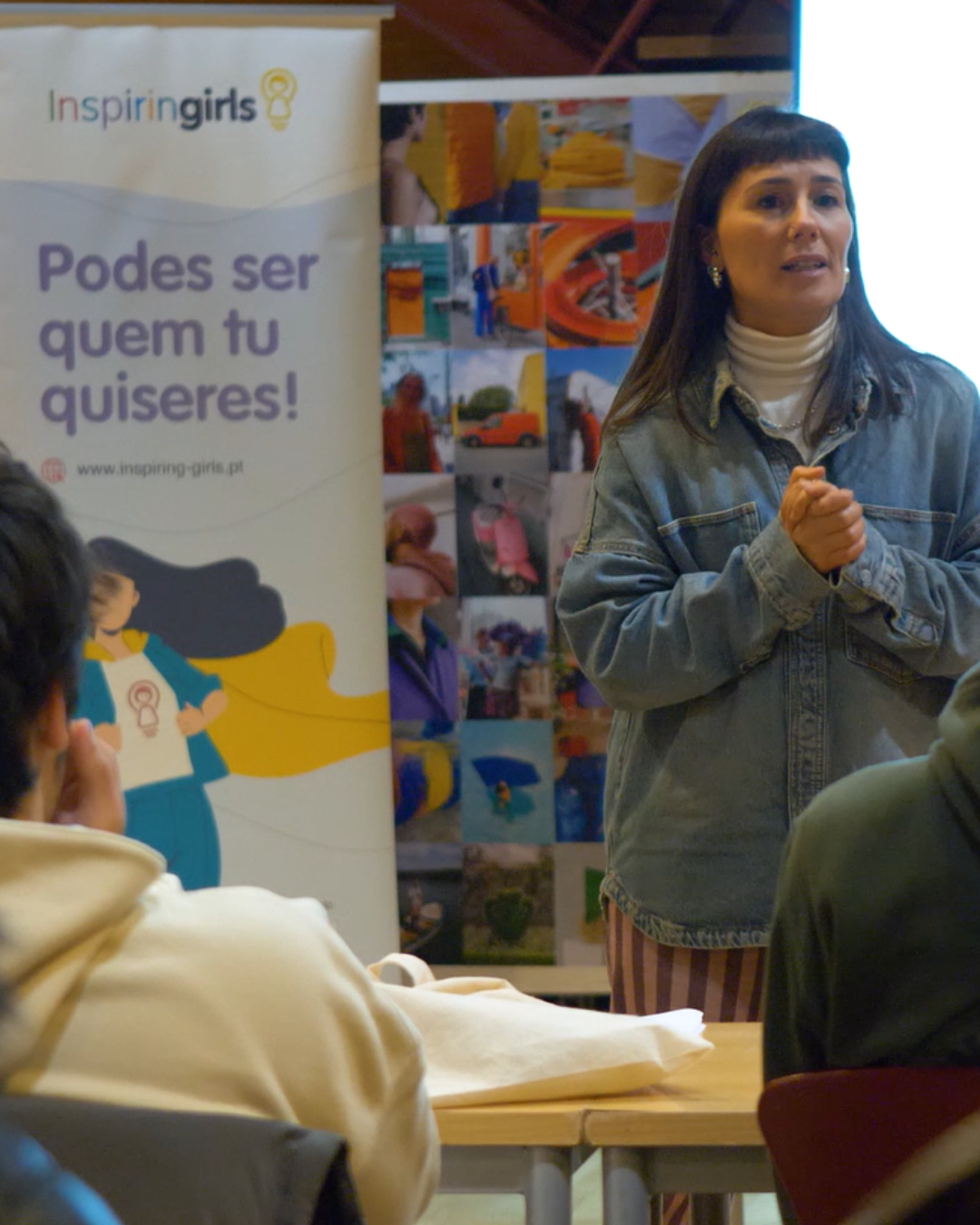 A woman is talking to a group of students. She is wearing a denim shirt and stripy trousers. She has her hands clasped together.