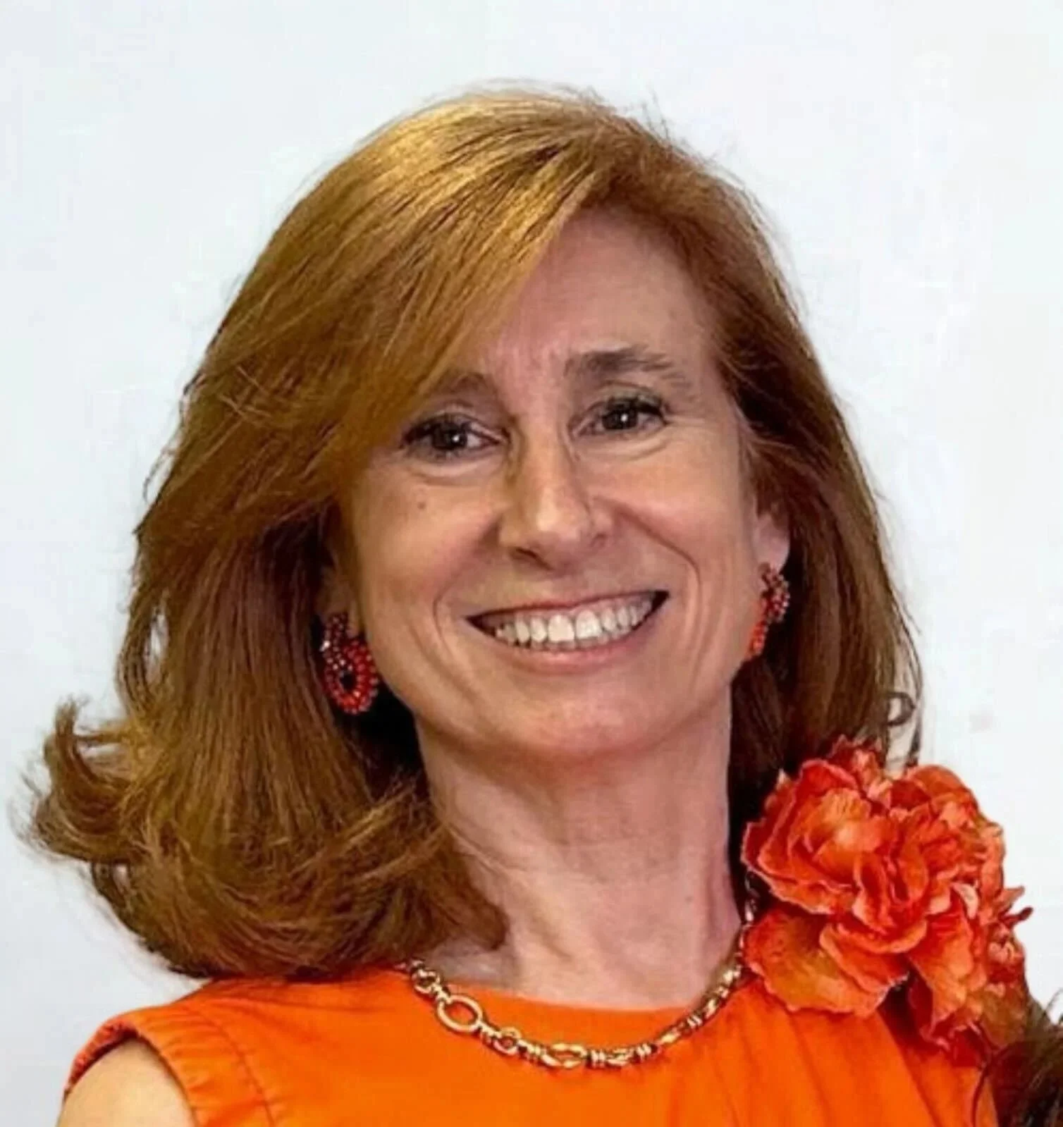 A woman with shoulder-length red hair smiling, wearing an orange top with an orange flower on right shoulder, against a white background.