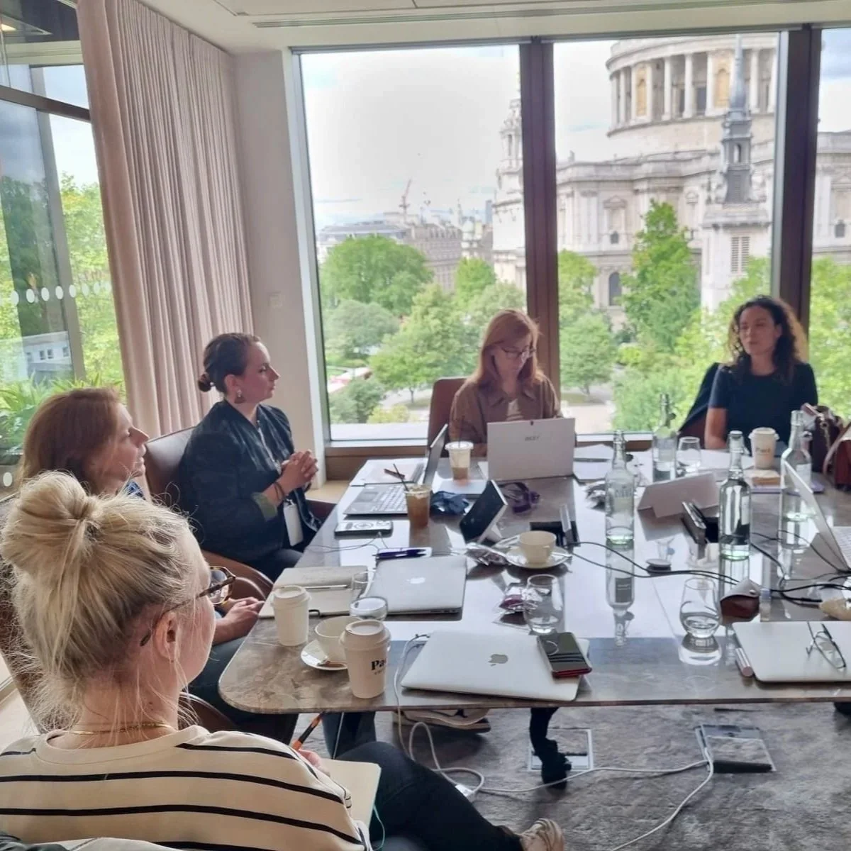 Meeting with five women sat around a table with laptops and views of London in the background.