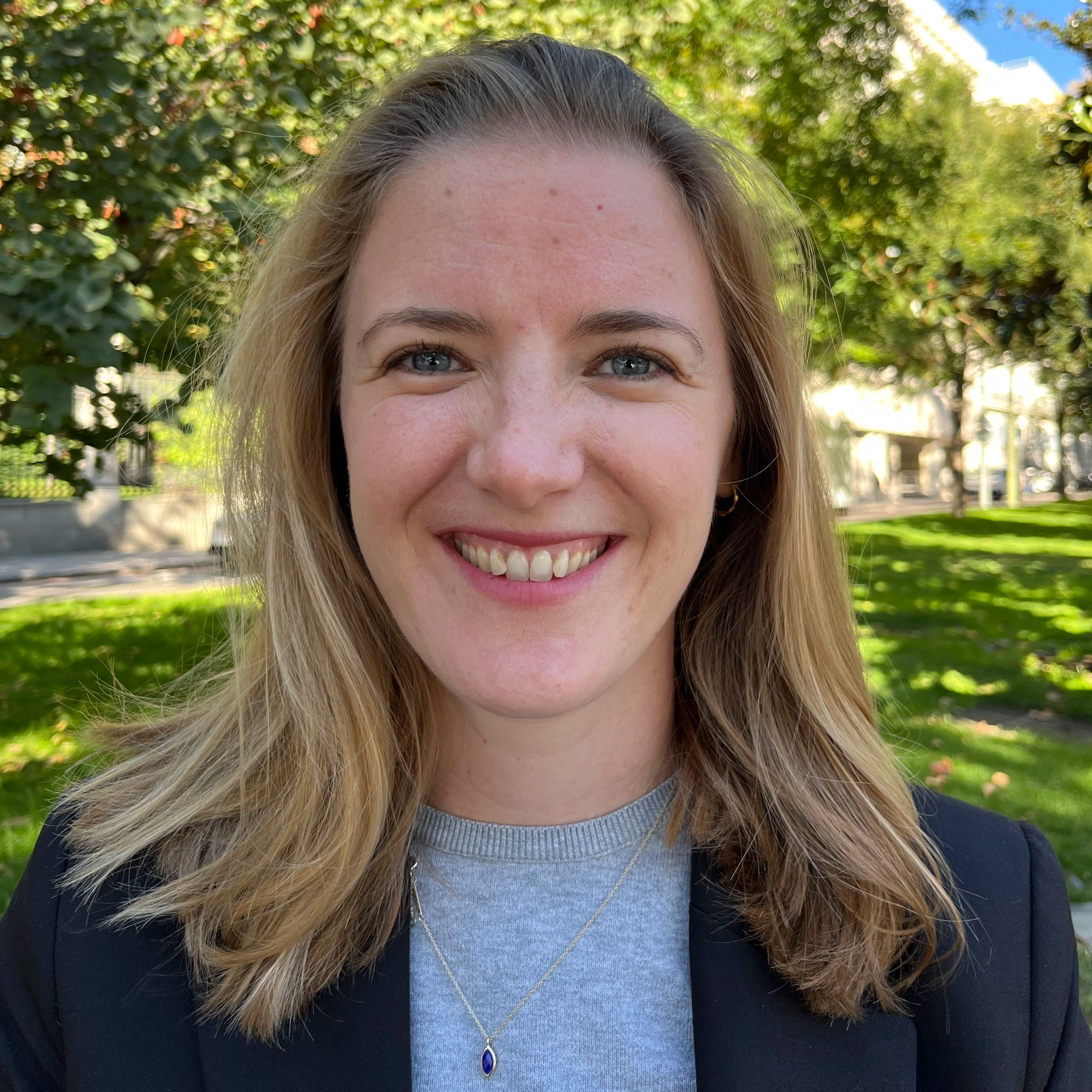 Woman smiling to camera, weating a grey jumper and black blazer, with trees in the background.