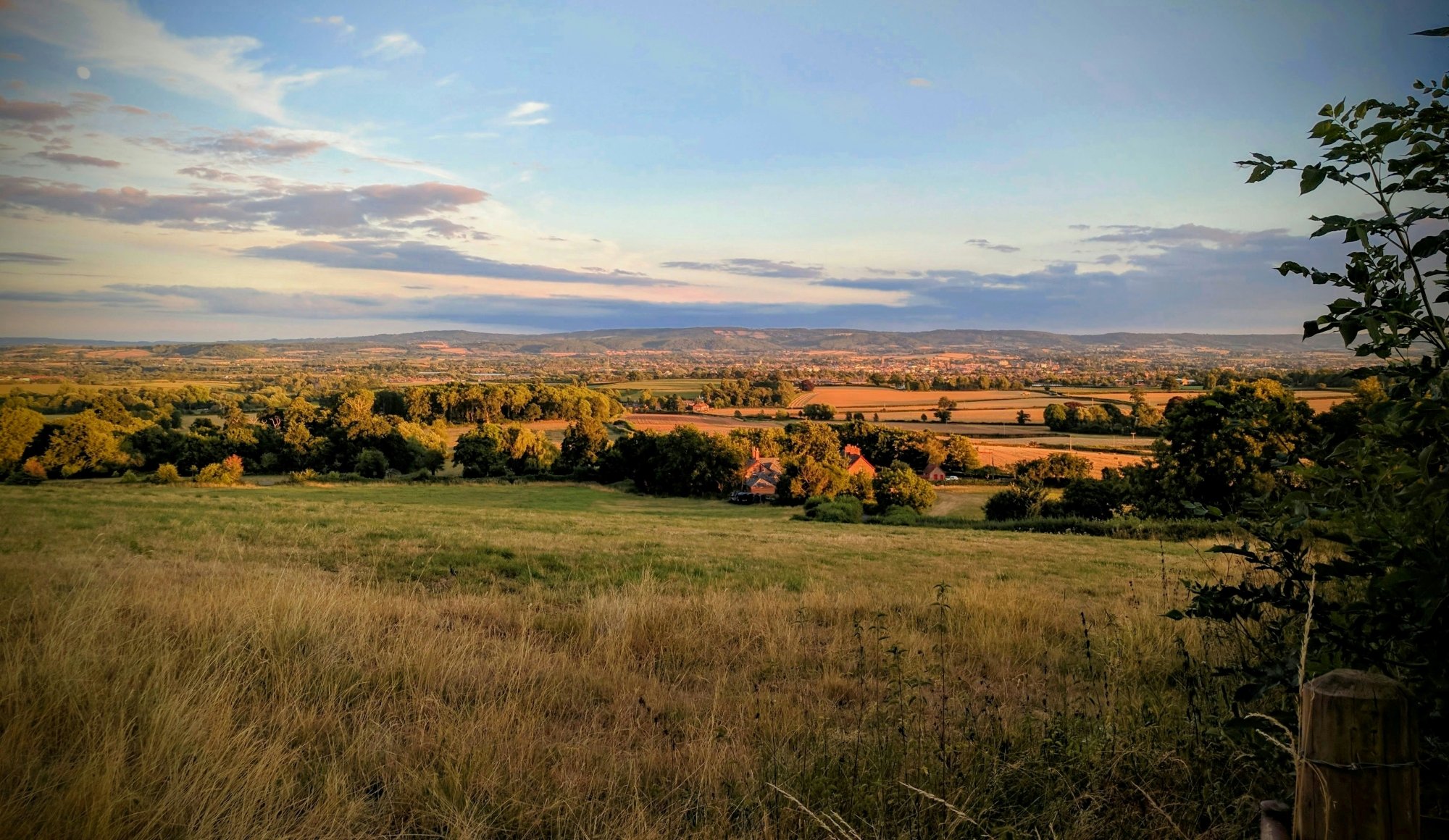 A scenic view of rolling fields, trees, and distant hills during sunset, with a clear sky and some scattered clouds, in Somerset, England.