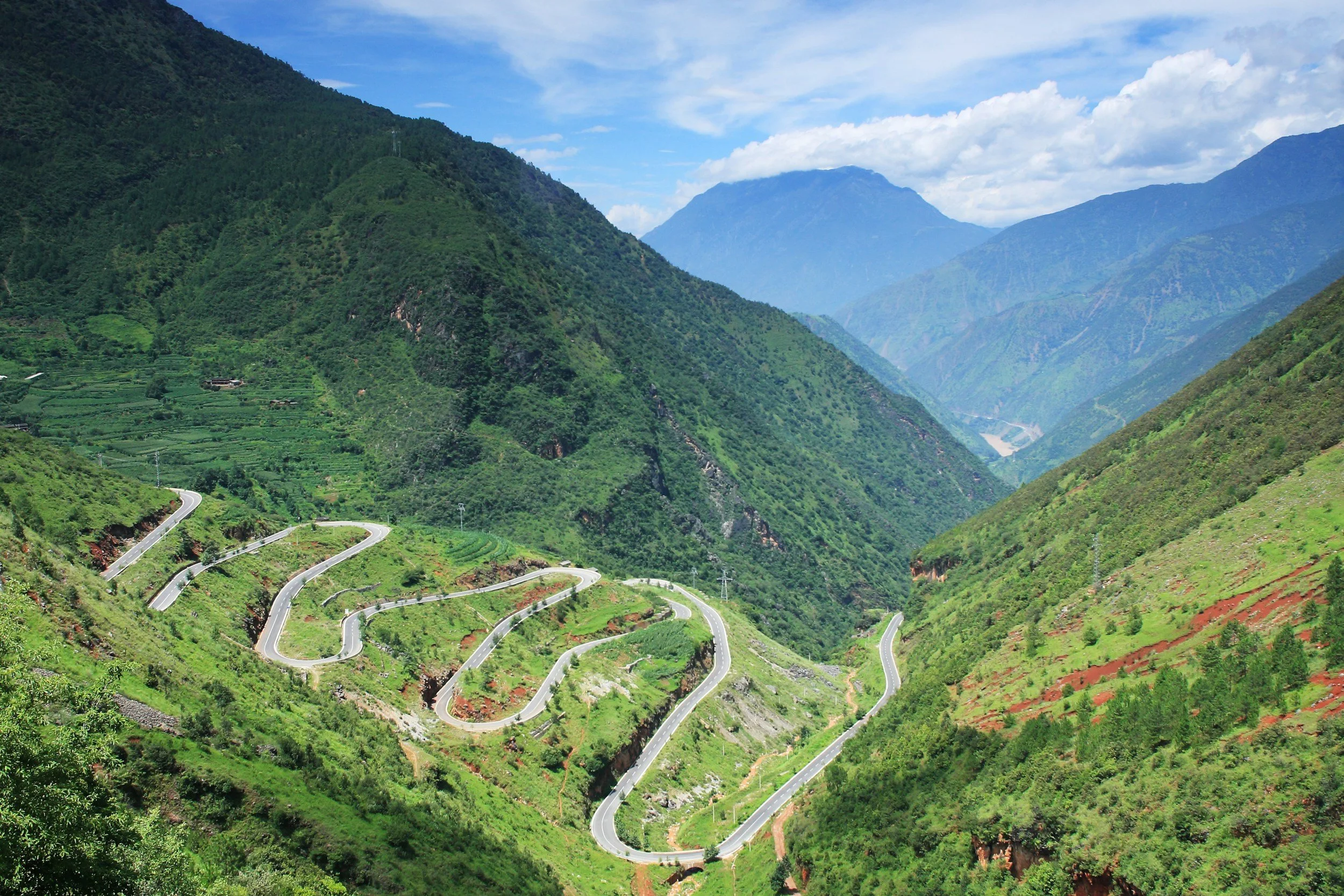 Winding mountain road through lush green hills and valleys with distant mountains in the background.