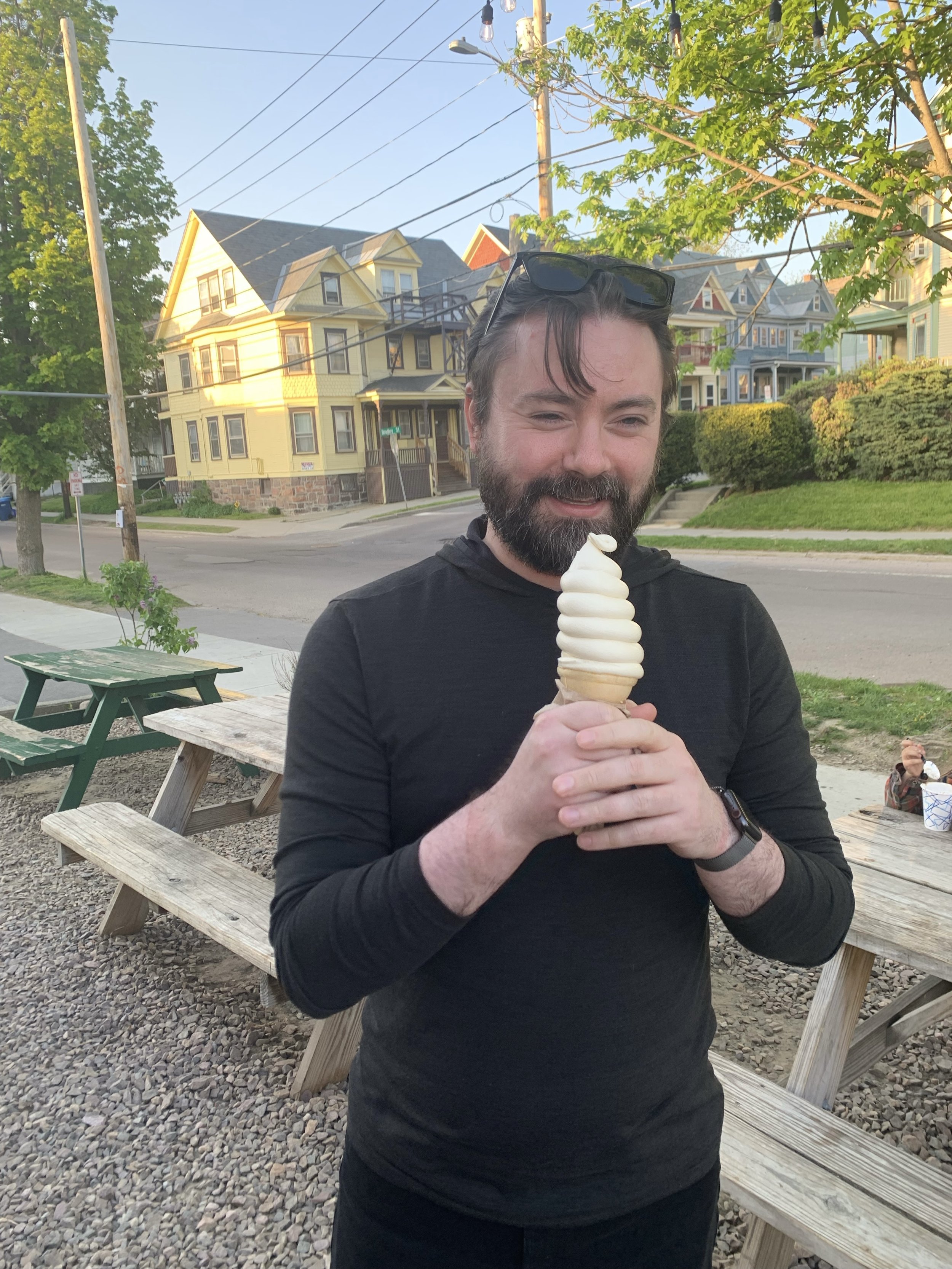 A man with a beard and glasses on his head holding a vanilla soft serve ice cream cone outdoors in a residential area with colorful houses and green trees.