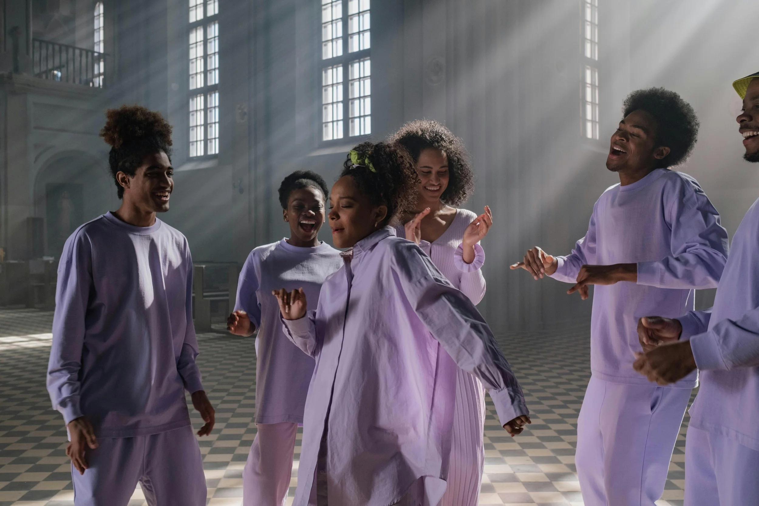 A group of people wearing white clothing are dancing indoors in a space with large windows and light streaming in.