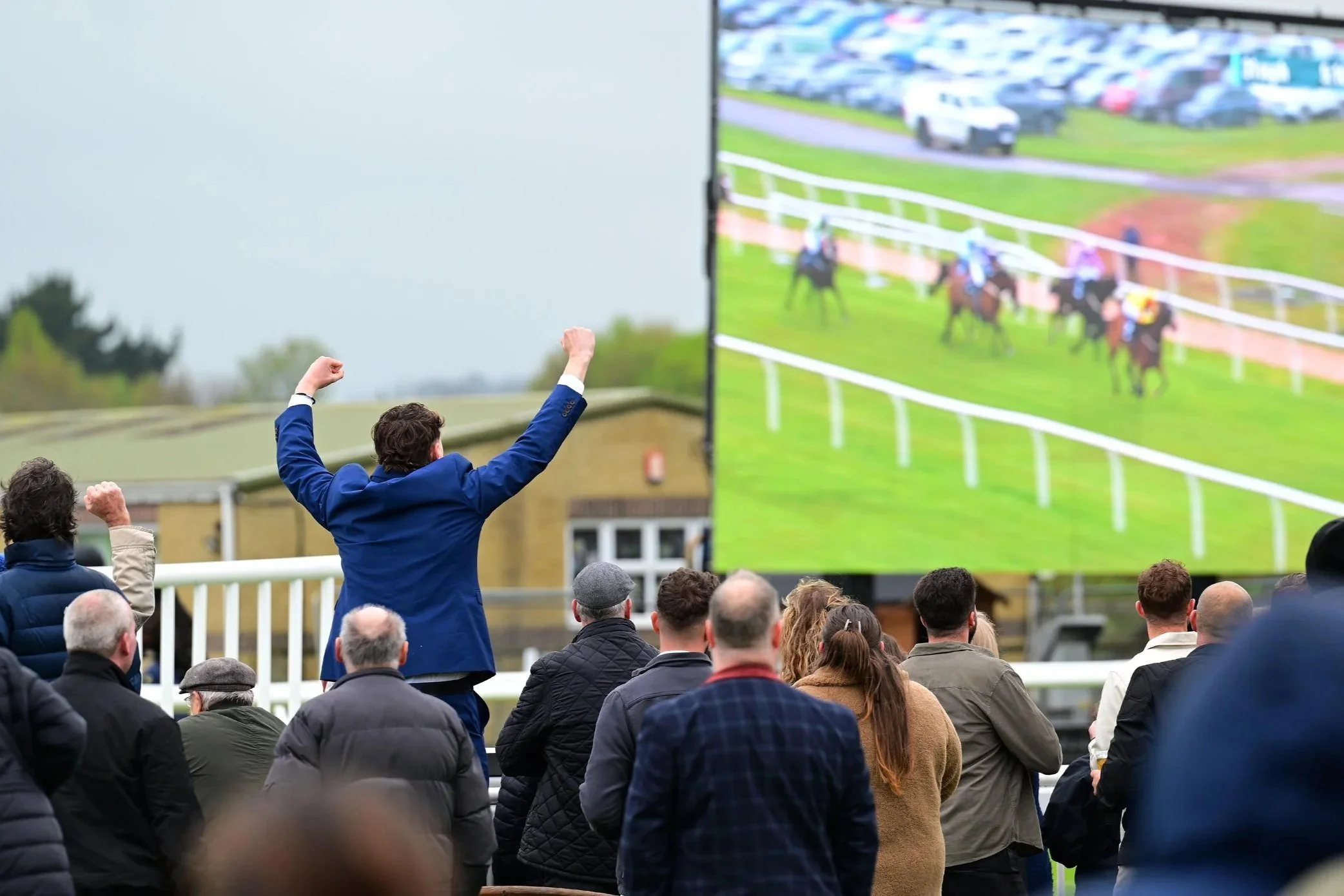 Crowd of people watching a horse race on a large screen at an outdoor event.