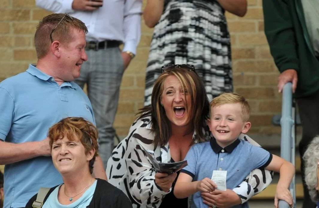 People laughing and smiling, including a woman with sunglasses on her head and children, at an outdoor event.