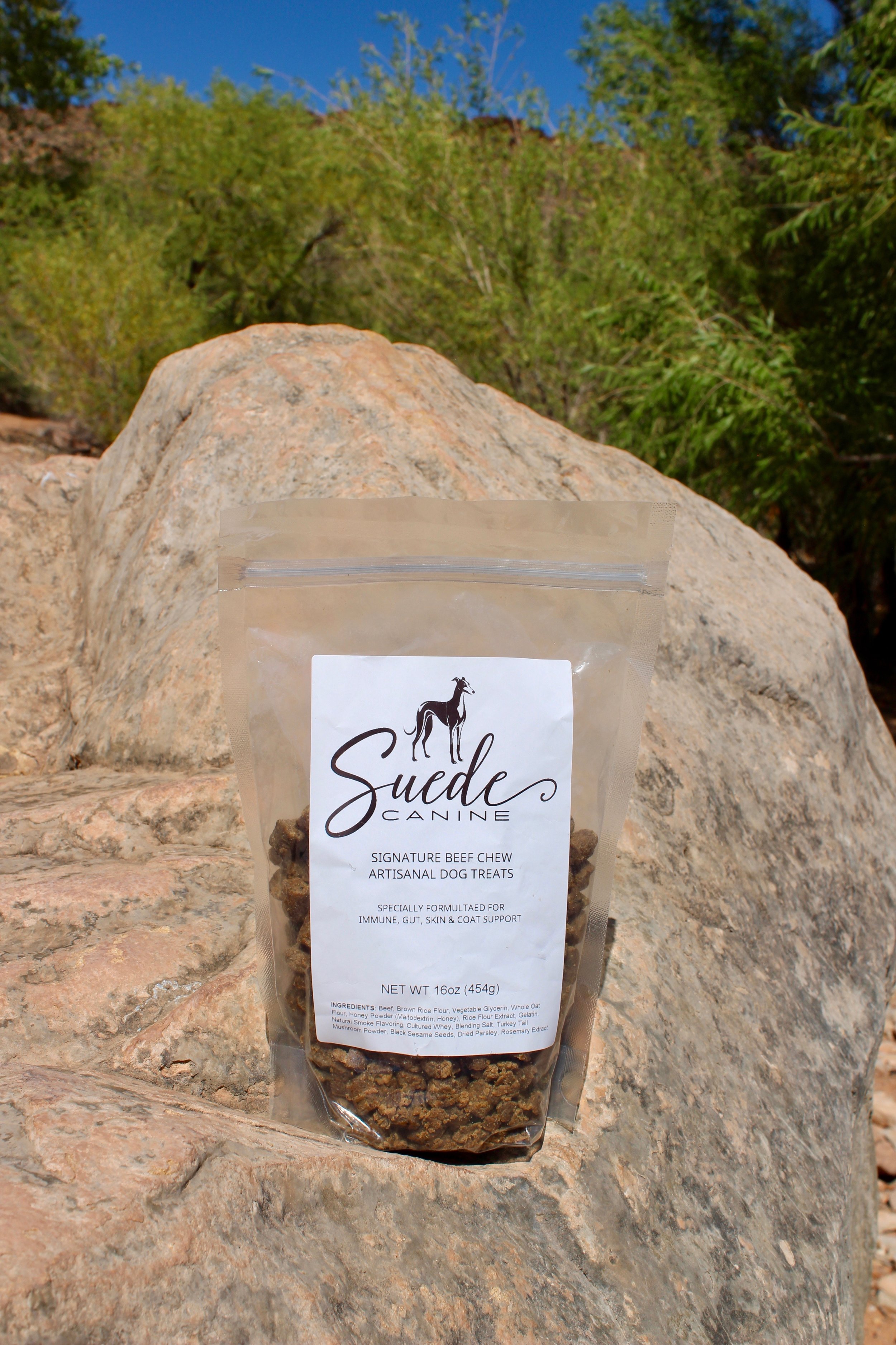 A clear plastic bag of dog treats labeled 'Suede Canine Signature Beef Chew' resting on a rock outdoors with green trees and a blue sky in the background.
