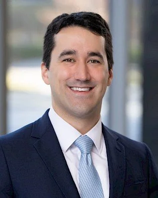 Professional man in a navy suit, light blue dress shirt, and tie smiling, with a blurred office background.