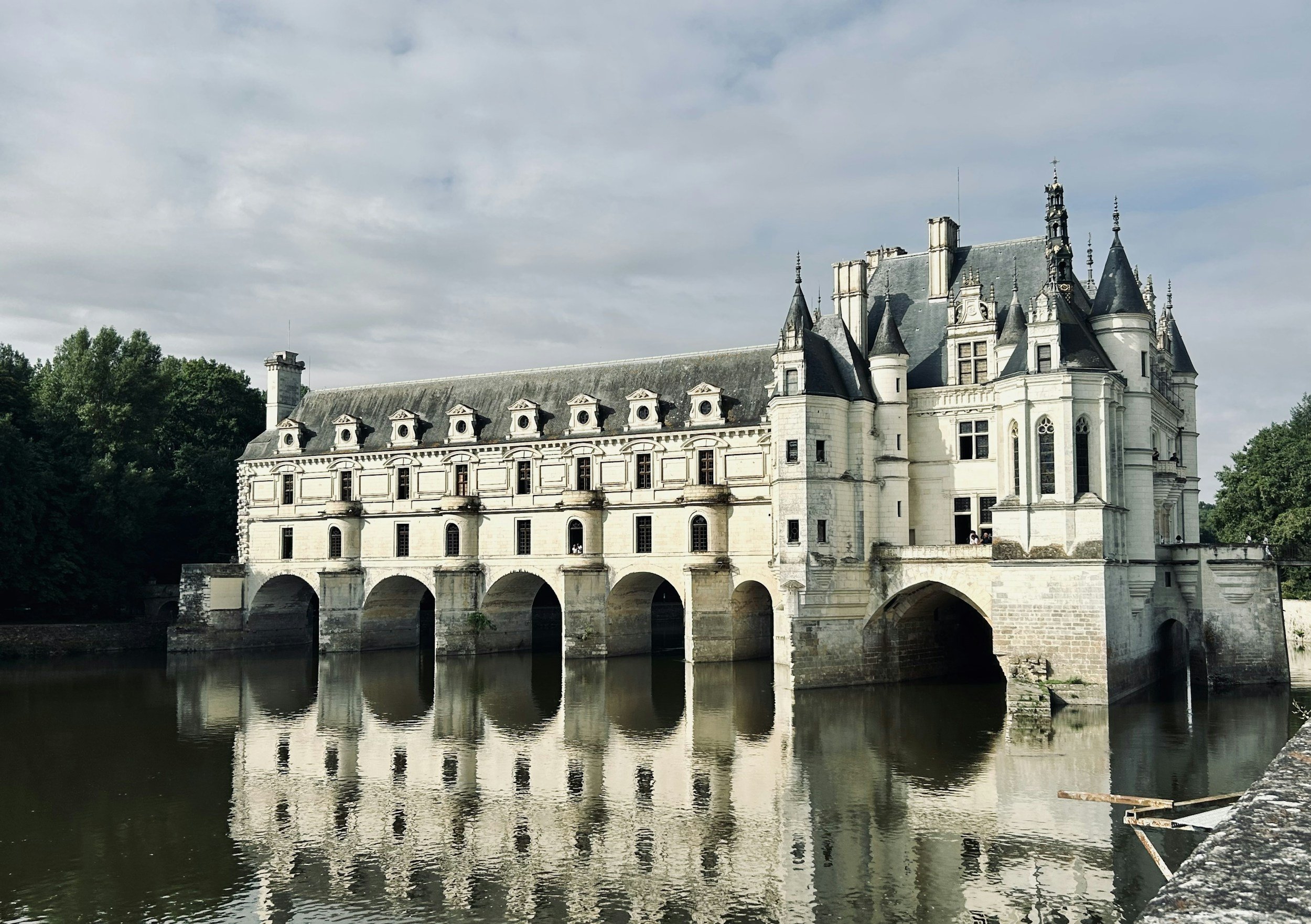 A historic castle-like building with multiple towers and pointed roofs alongside a river with reflections, surrounded by greenery under a cloudy sky in the Loire Valley (Chateau Chenonceau)