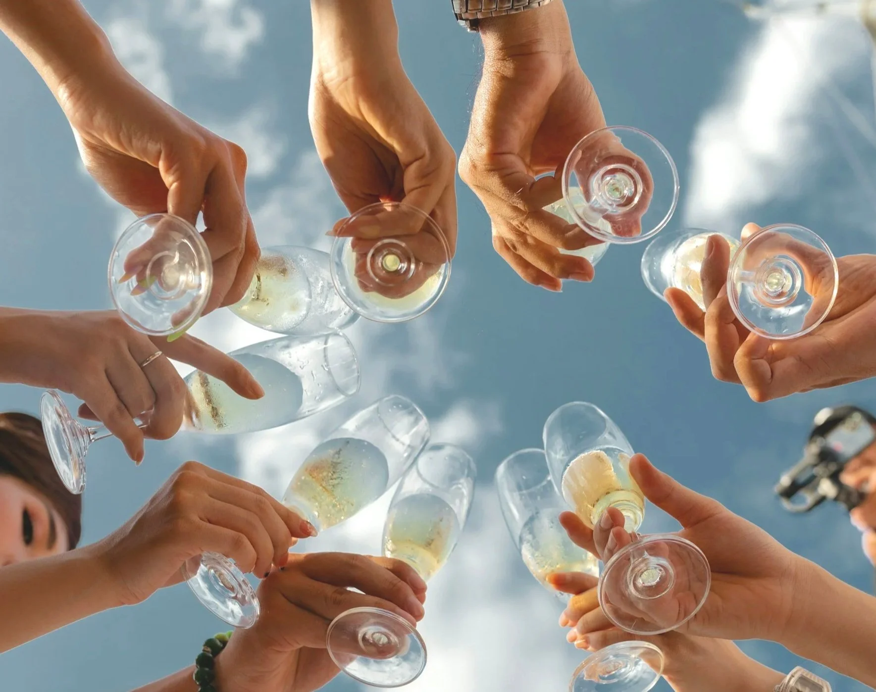 Multiple people clinking champagne glasses together in a toast outdoors against a blue sky with clouds.