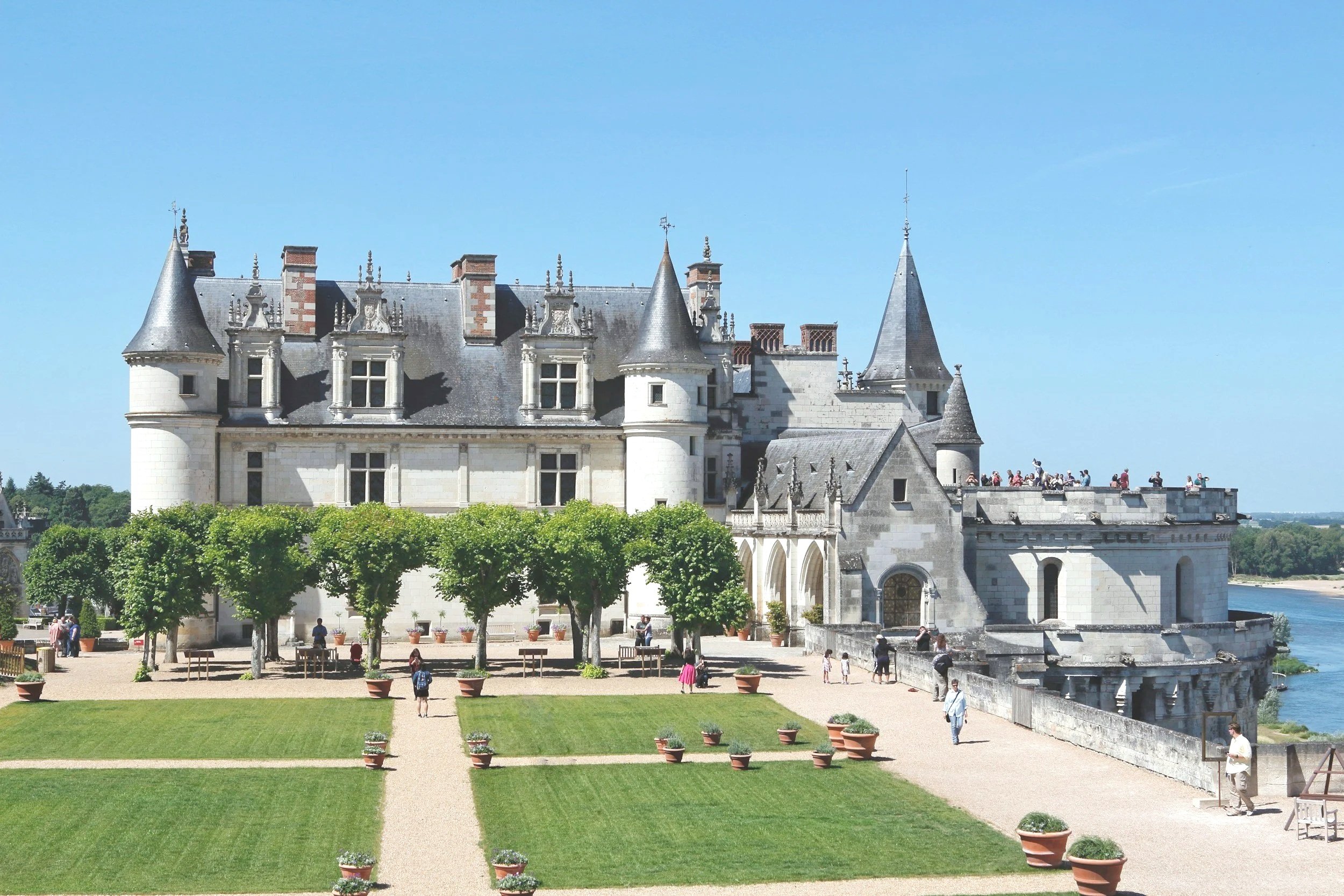 A majestic castle with turrets and chimneys above stone walls, overlooking a river, with a well-manicured garden with paths and potted plants in the foreground, and many tourists exploring the grounds in the Loire Valley (Chateau d'Amboise)