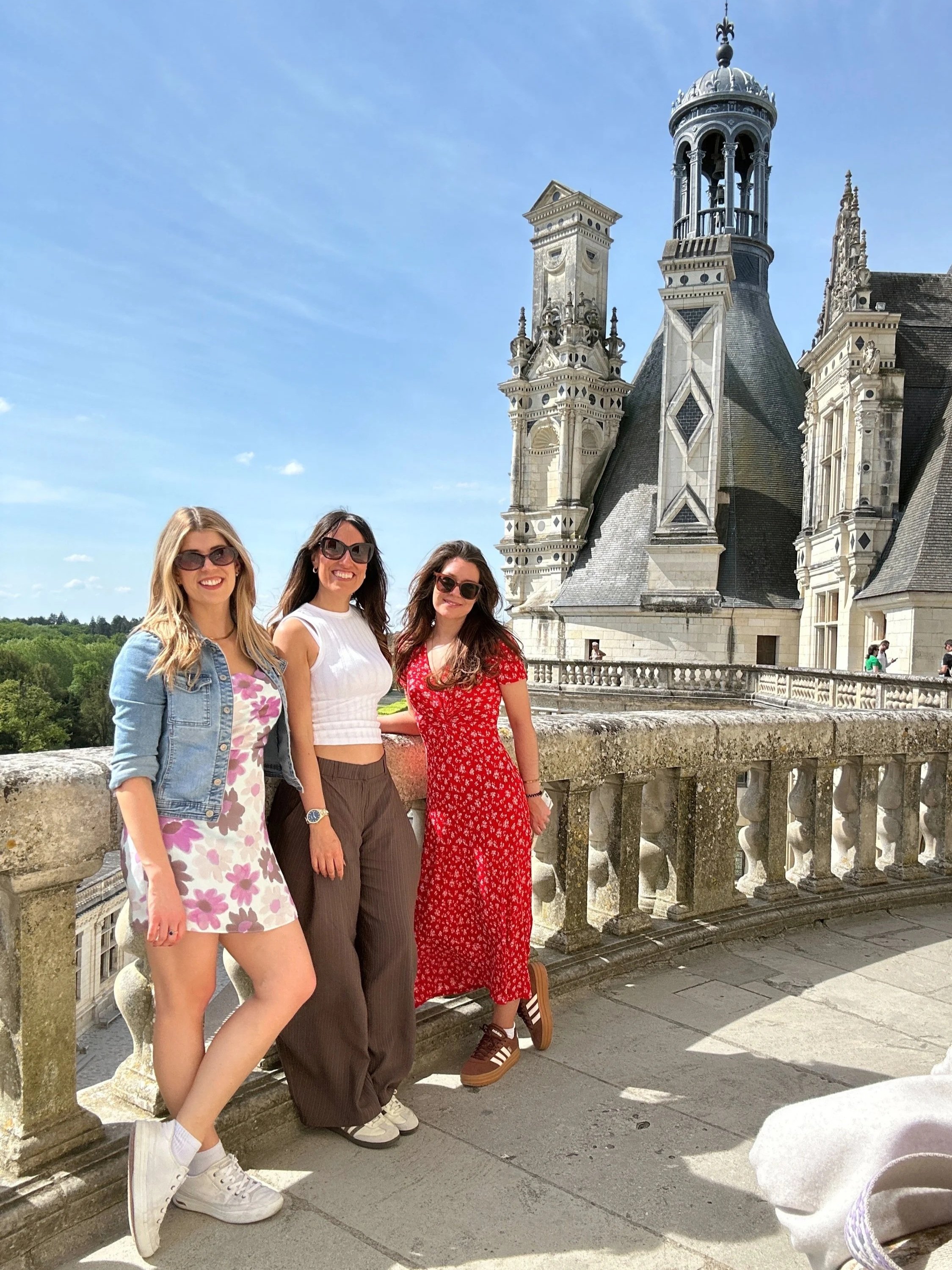 Three women smiling and standing together on a balcony of a historic building with ornate towers and a clear blue sky behind them. Paris, Champagne, and The Loire Valley wine retreat for women
