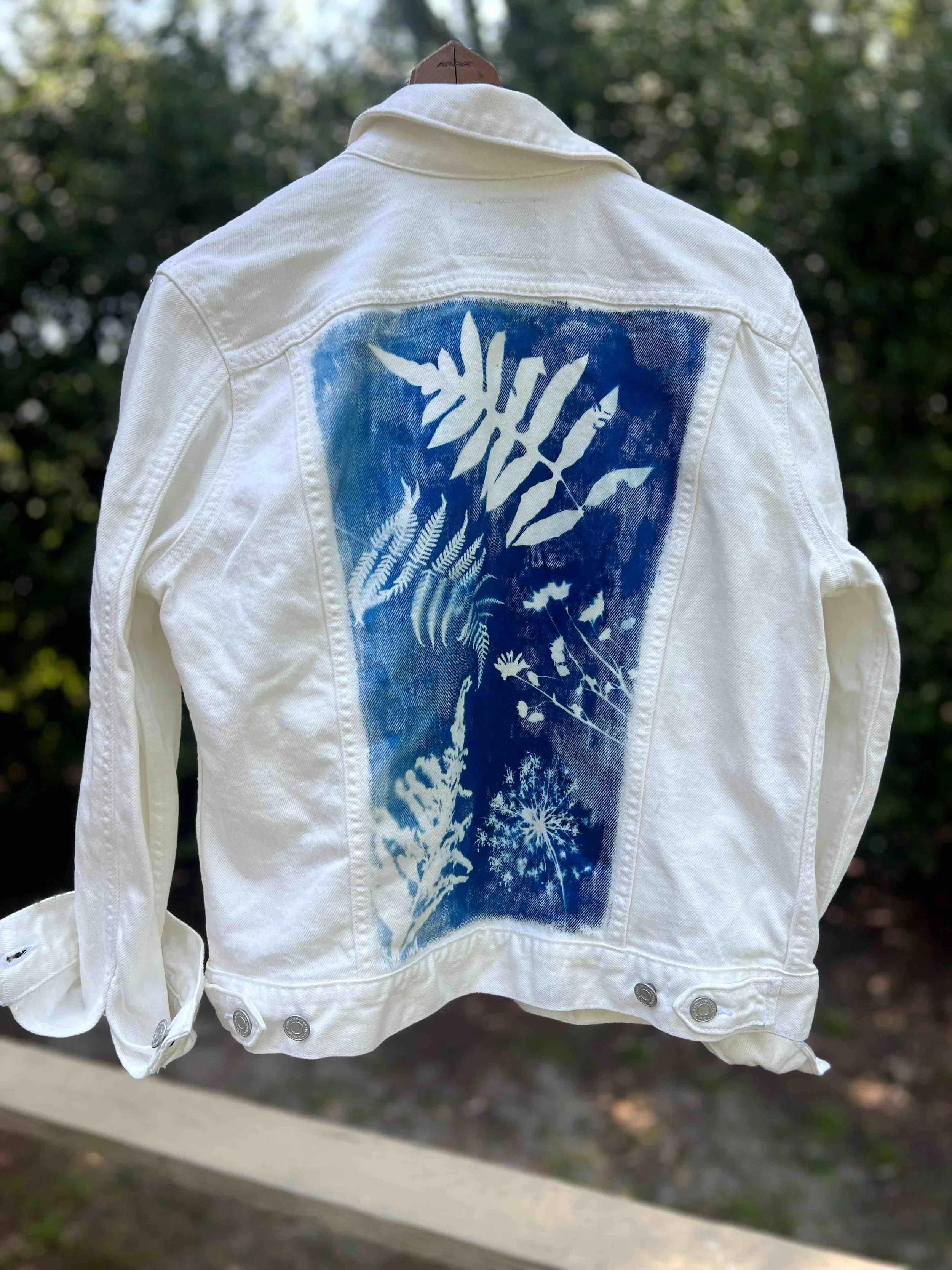 White denim jacket hanging outdoors, with a blue graphic cyanotype design featuring leaves, flowers, and a star-shaped pattern on the back.