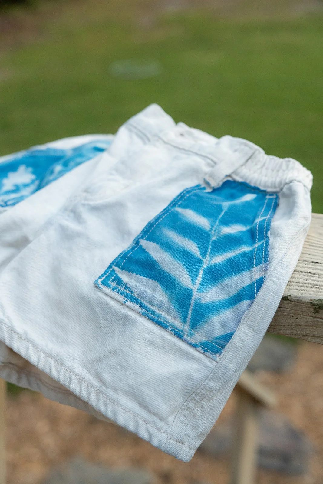 A pair of white and blue swim trunks with a leaf pattern on the pocket laid on a wooden surface outdoors.