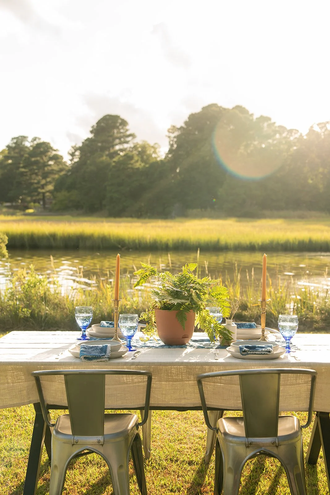 A table set outdoors near a lake with a lush green background, featuring a potted plant, blue glasses of water, blue napkins, rice, and candlesticks with unlit candles, illuminated by warm sunlight.