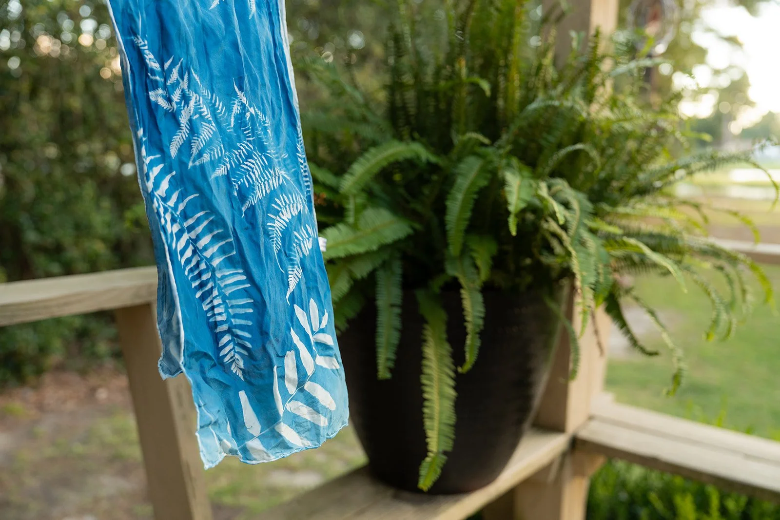 A blue fabric with white leaf patterns hanging from a wooden railing next to a large potted fern plant on a wooden shelf.