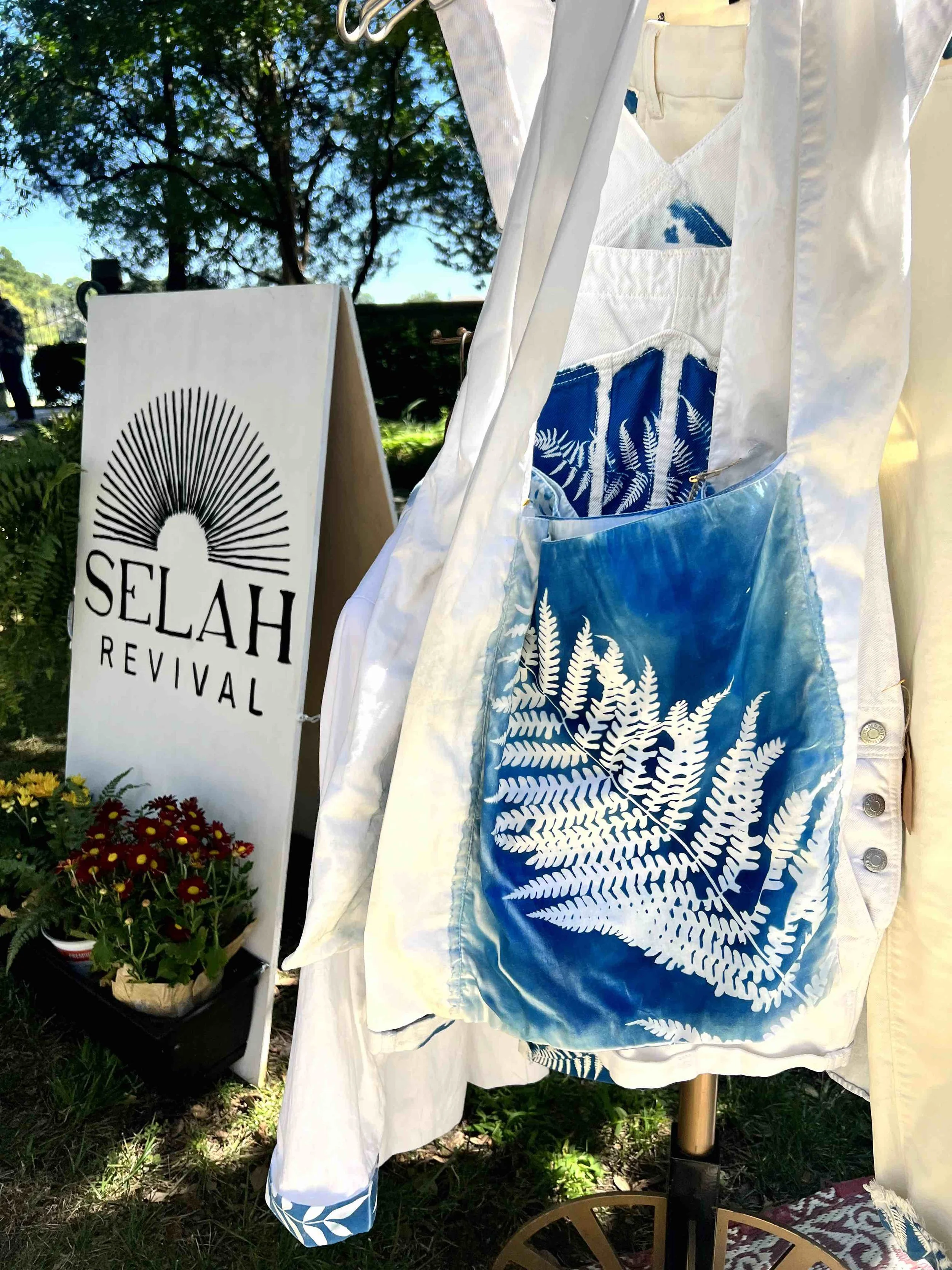 Outdoor scene with a white stand-up sign that reads 'Selah Revival'. Part of a display with white and blue fabric featuring white fern and leaf patterns, hanging from a clothing rack. Trees and a blue sky can be seen in the background.