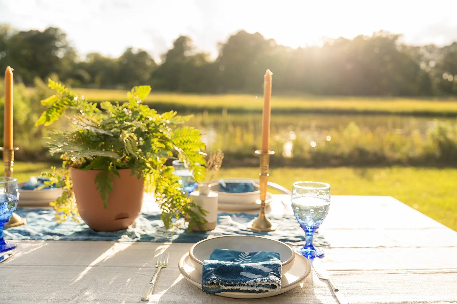 Outdoor dining table set with plates, drinking glasses, candles, and a potted plant, illuminated by sunset with a river and trees in the background.