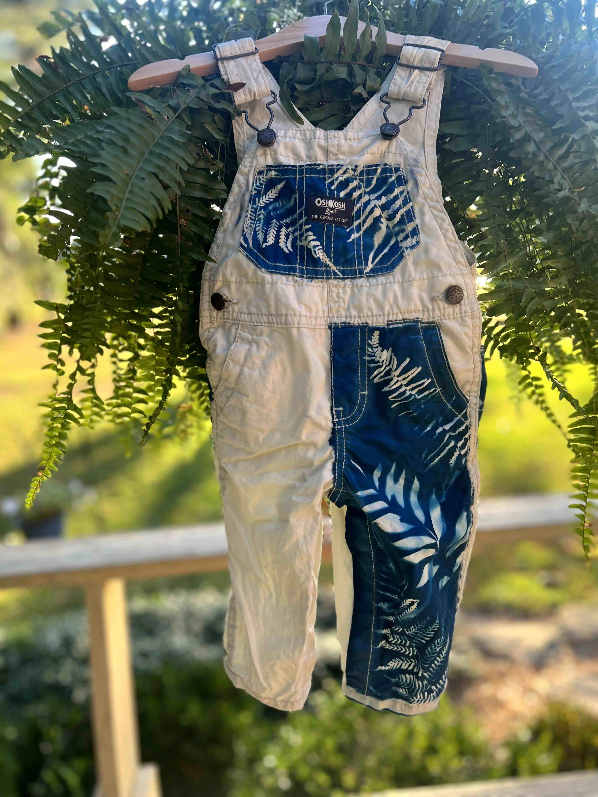 Two pairs of denim overalls, one in light wash with a leaf pattern on the front, and the other in darker wash with a similar leaf pattern, hanging on a wooden hanger from a leafy tree branch with a blurred background of green foliage.