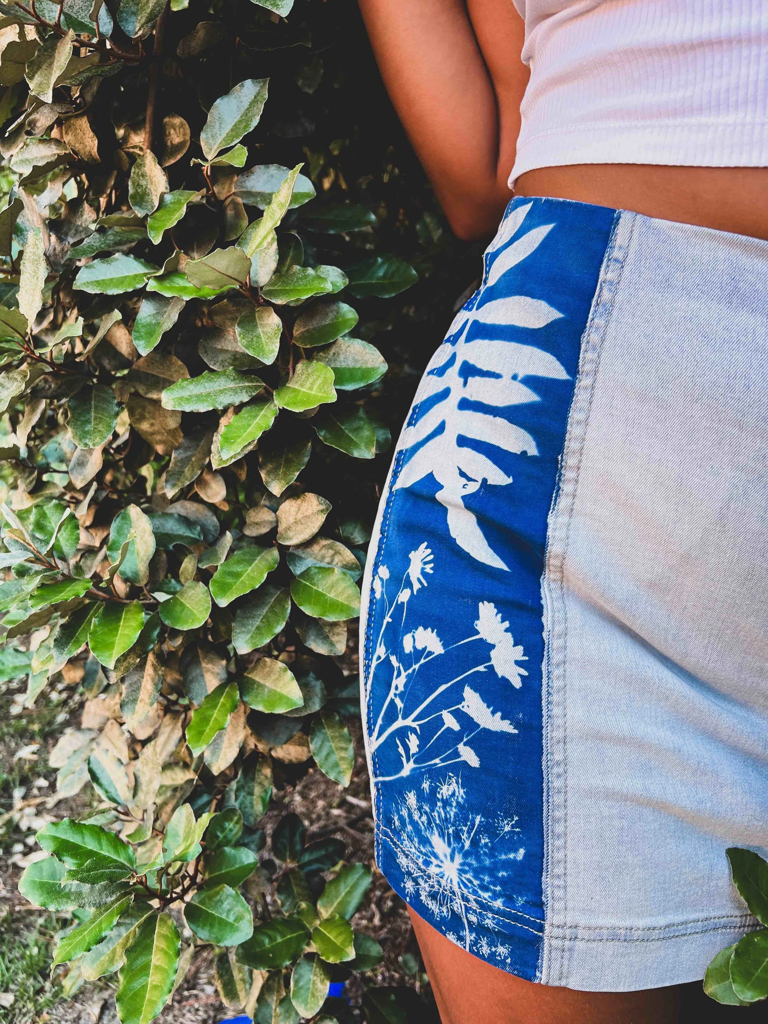 Close-up of a person's lower torso wearing blue and white floral patterned shorts and a white crop top, standing next to a hedge of green leaves.
