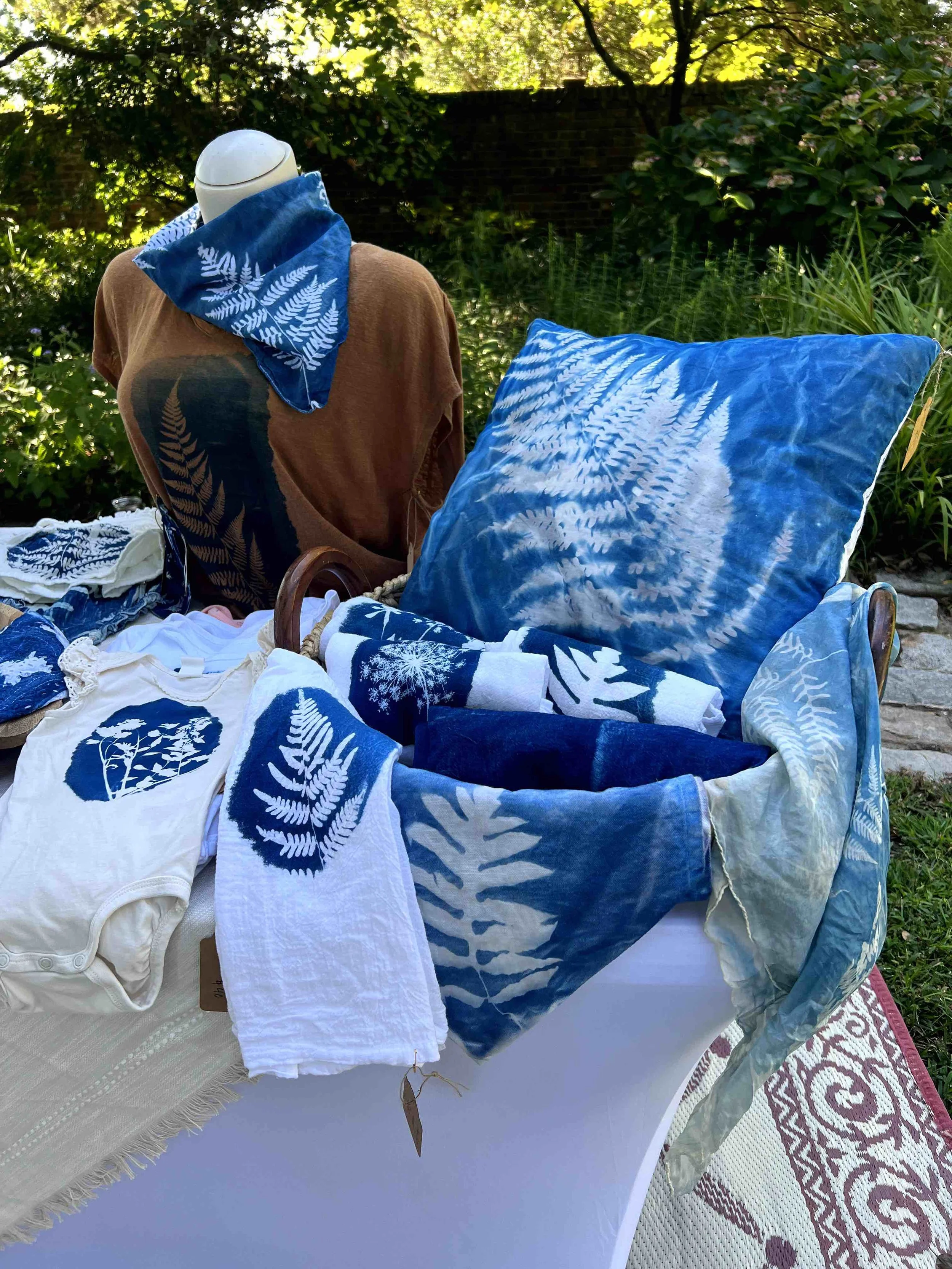 Outdoor display of blue and white fabric towels and linens with fern and leaf patterns on a bed or table, set in a garden with greenery and trees in the background.