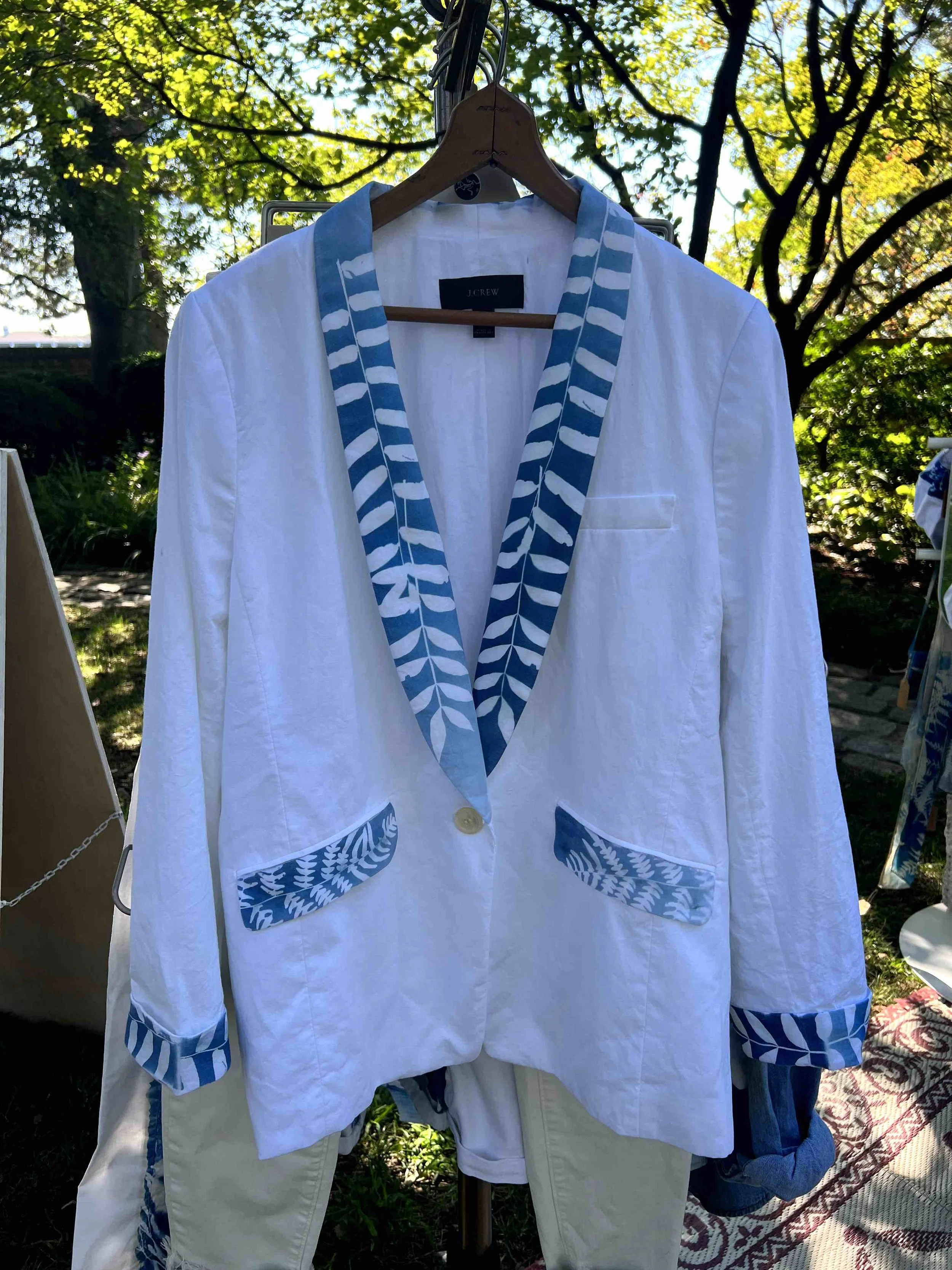White blazer with blue and white leaf-patterned lapel, pockets, and cuffs hanging on a wooden hanger outdoors surrounded by trees and greenery.