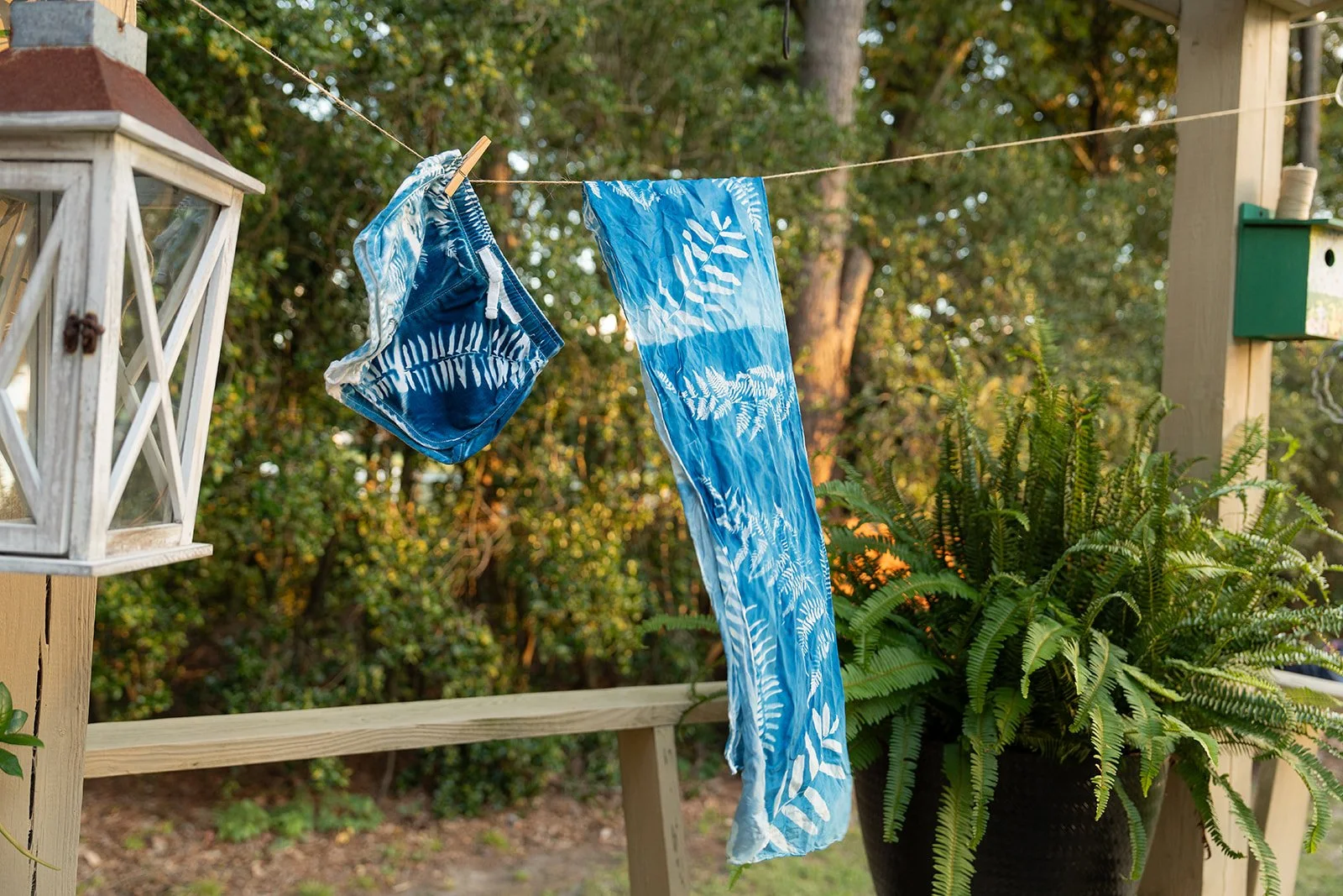A clothesline with blue and white patterned swim cyantoype shorts and a matching towel hanging on it outdoors, with a wooden deck, a lantern, a potted fern, and trees in the background during daytime.