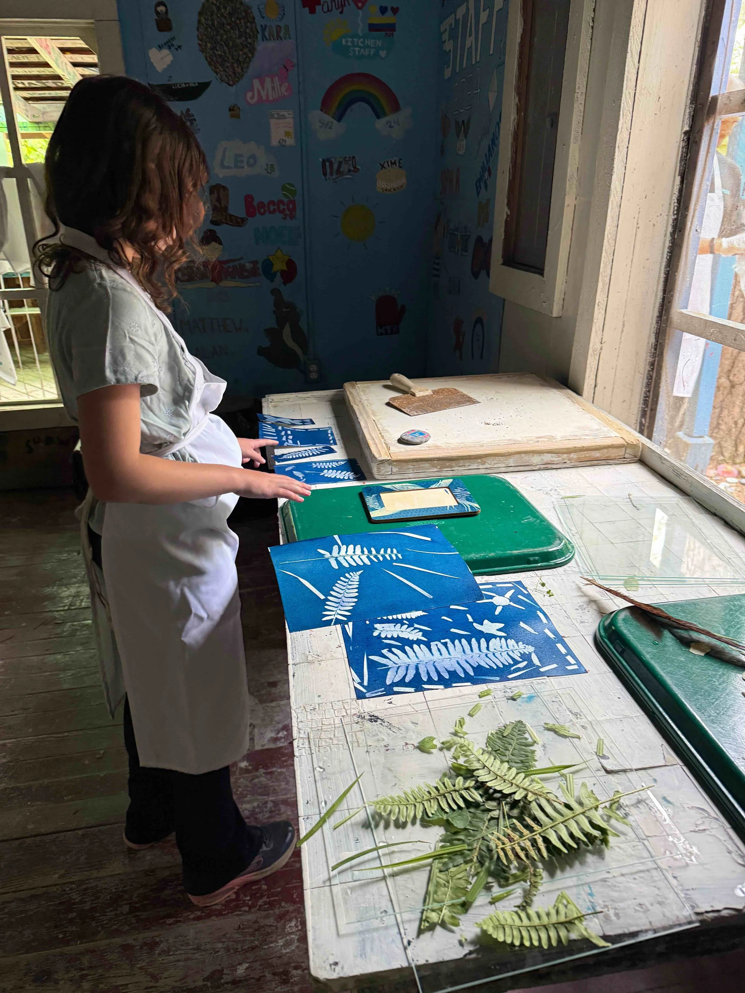 A young girl wearing a white apron and mask stands at a table, doing cyanotype printmaking with blue paper and greenery. The table has green trays, a cutting tool, and cut leaves for printmaking.