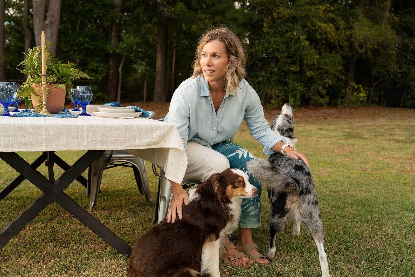 Woman sitting outdoors at a table with two dogs, one on her lap and one standing beside her. The table is set with plates, glasses, and a flower arrangement. The setting appears to be a backyard or park with trees in the background.