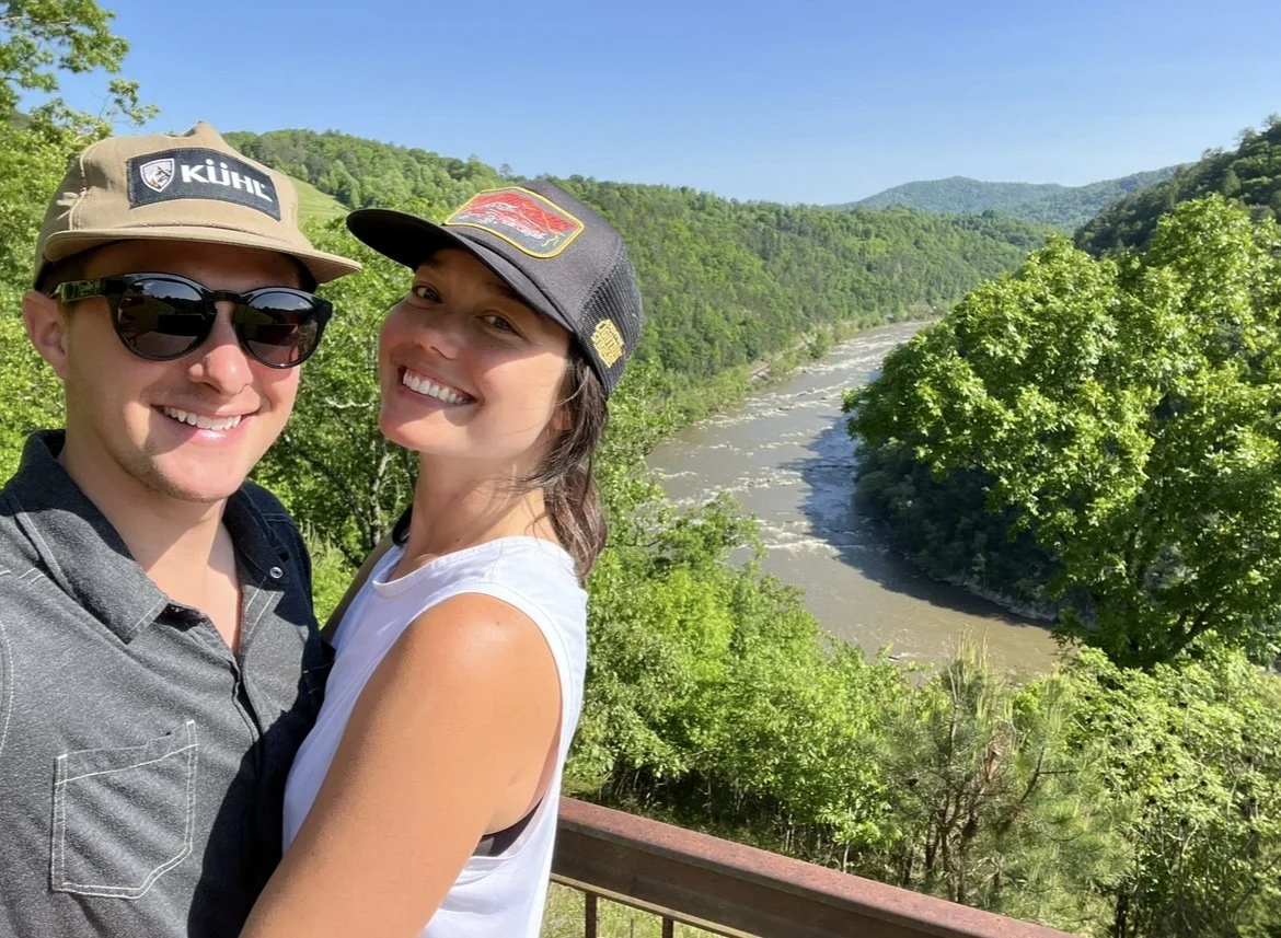 A smiling couple taking a selfie outdoors near a river with lush green trees and mountains in the background.