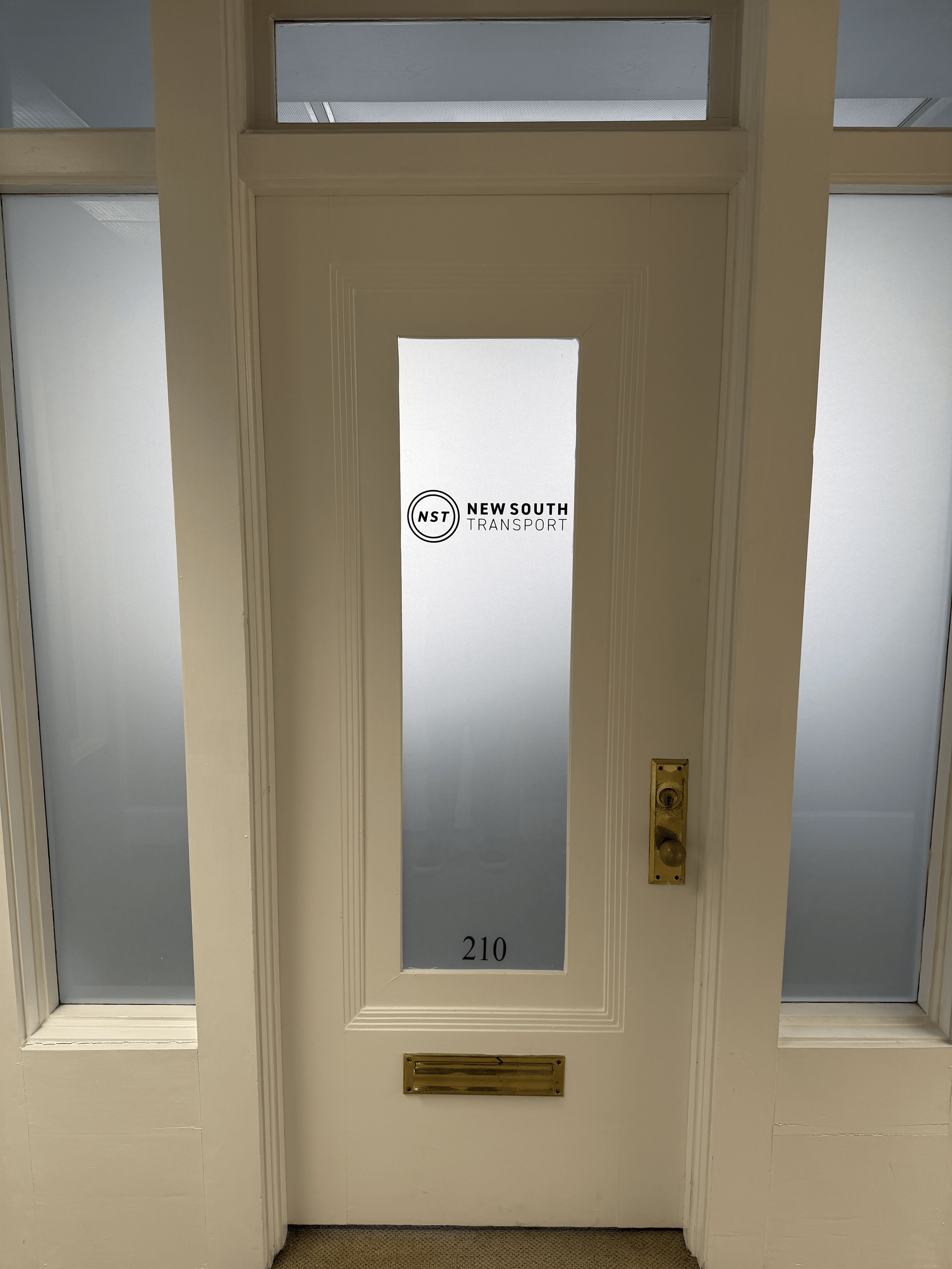 White door with a glass panel in the center labeled '210', locked door handle, and a brass mail slot, with frosted glass panels on either side, and a sign with the New South Transport logo on the glass panel.