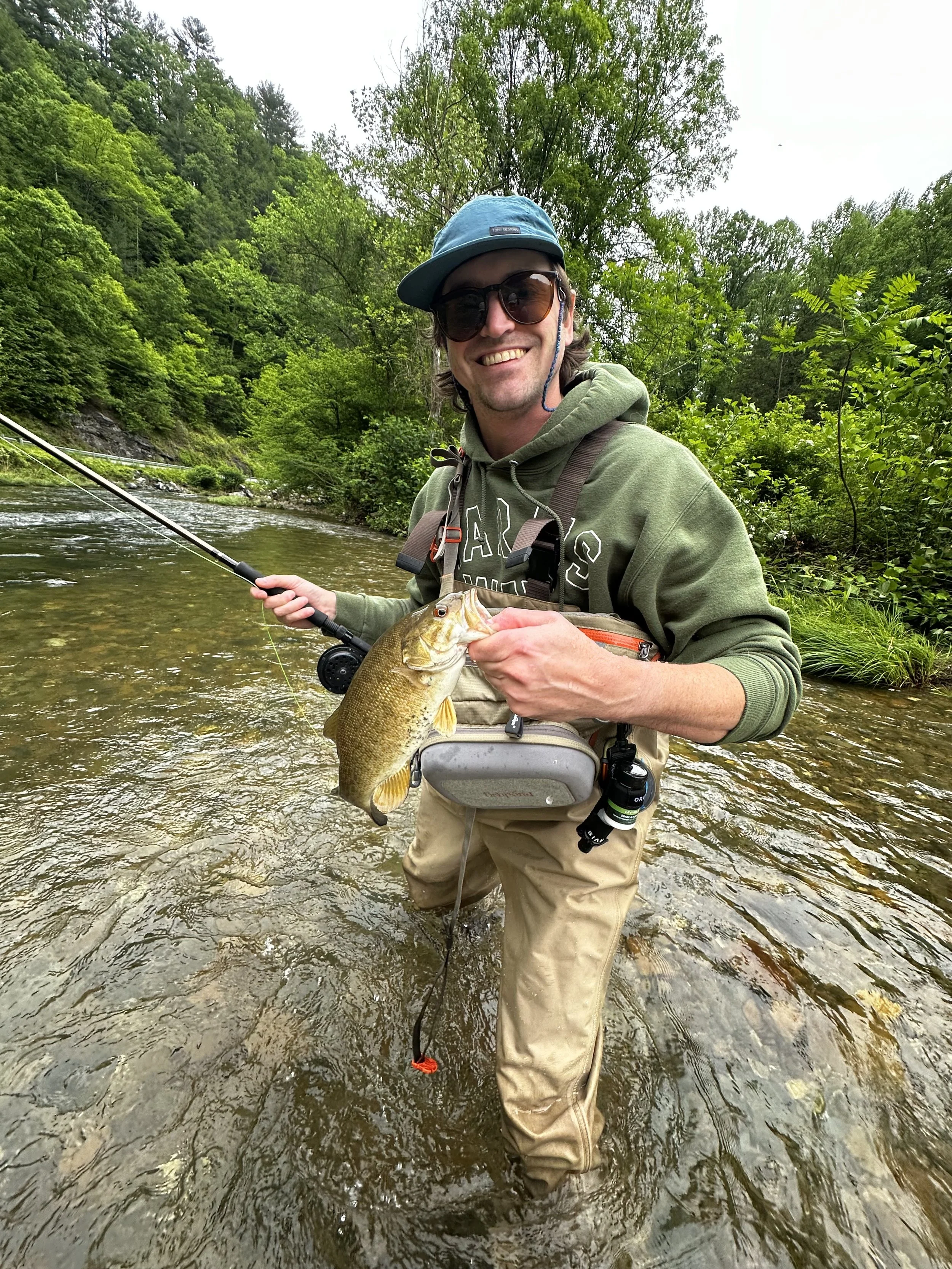 A man wearing sunglasses, a blue cap, a green hoodie, and khaki waders stands in a shallow river, smiling while holding a fishing rod in one hand and a caught fish in the other, surrounded by green trees and a cloudy sky.