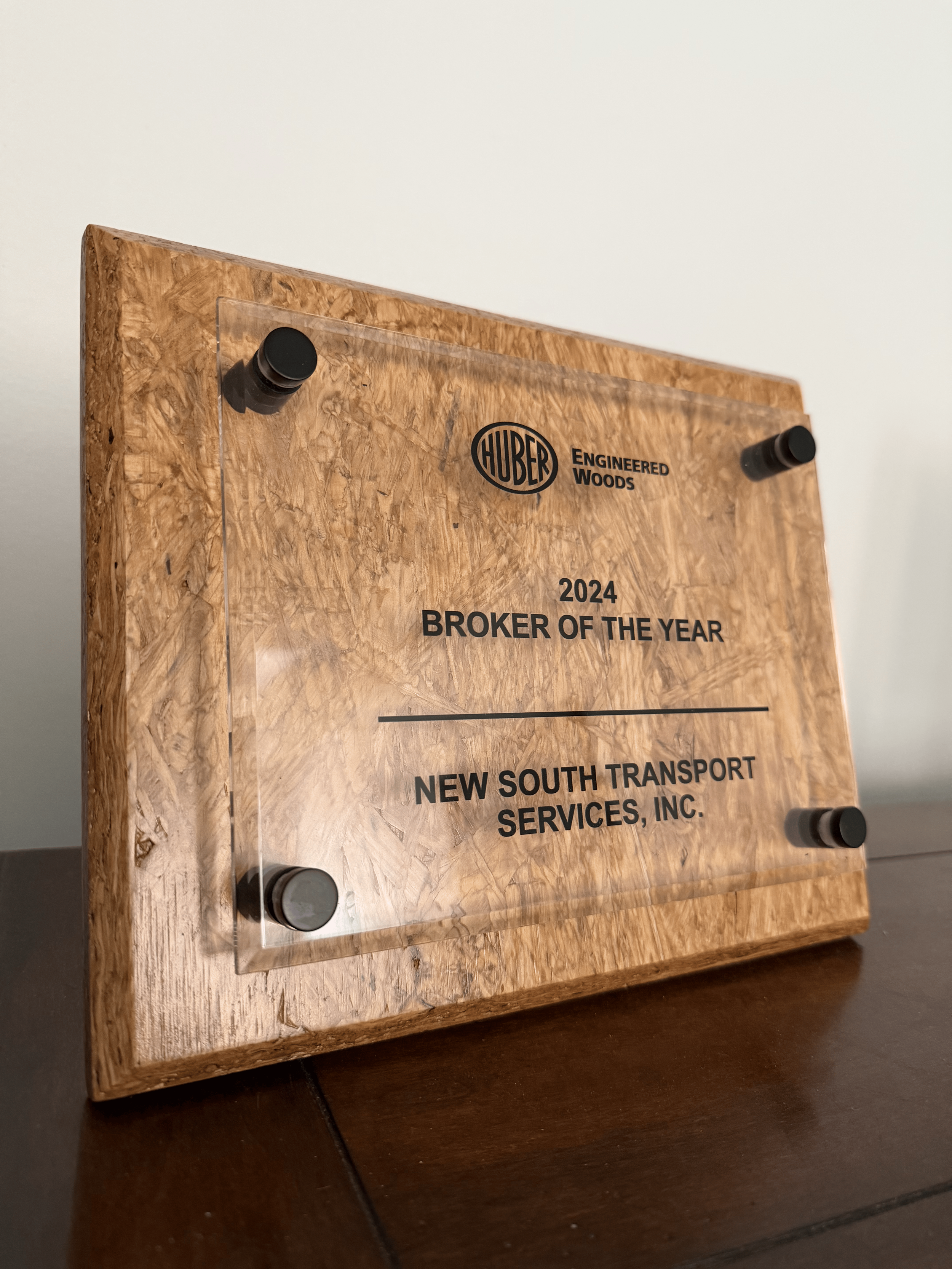 Wooden plaque with a transparent acrylic cover, mounted on a wall with four black standoffs. The plaque has the HUBER logo, the text "Engineered Woods," and the award information: "2024 Broker of the Year, New South Transport Services, Inc."
