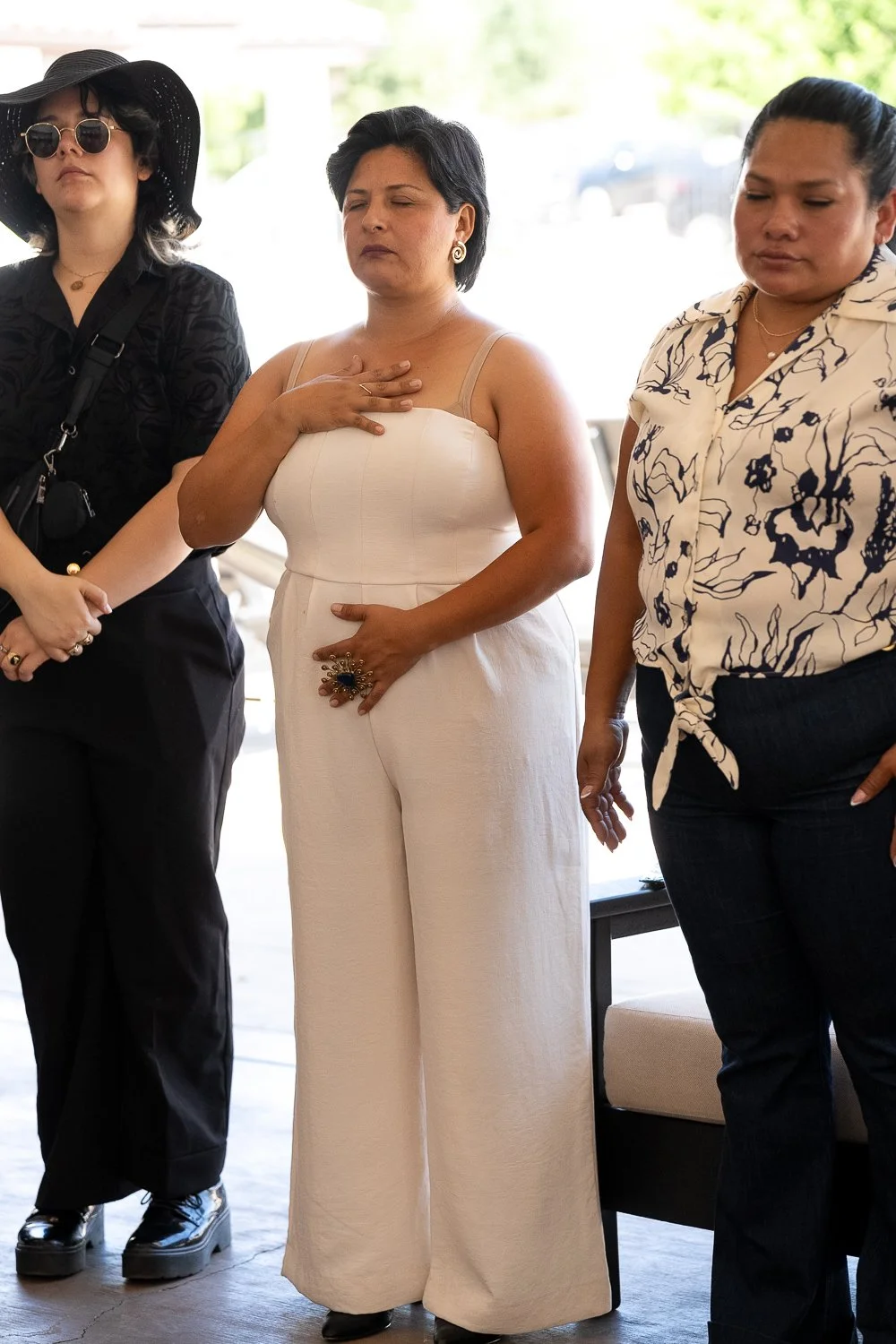 Three women standing with eyes closed, one with hand over her heart, in a moment of silence or prayer inside a well-lit room.