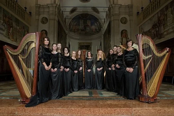 Un coro di donne in abiti neri, in piedi tra due grandi arpe, in una chiesa o un edificio storico con decorazioni religiose.
