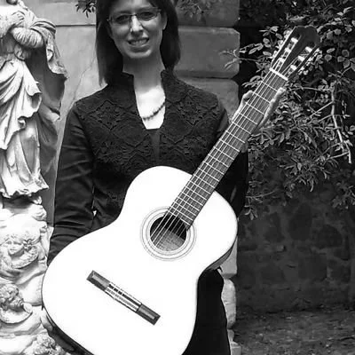 Una donna con capelli castani e occhiali, che tiene una chitarra classica di fronte a uno sfondo di alberi e sculture in pietra.