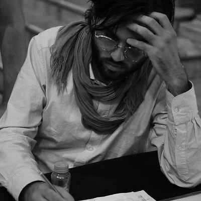 Persona seduta, con mano sulla fronte, che indossa occhiali, laicata un cappotto chiaro, con una sciarpa, davanti a un tavolo con una bottiglia e dei fogli.