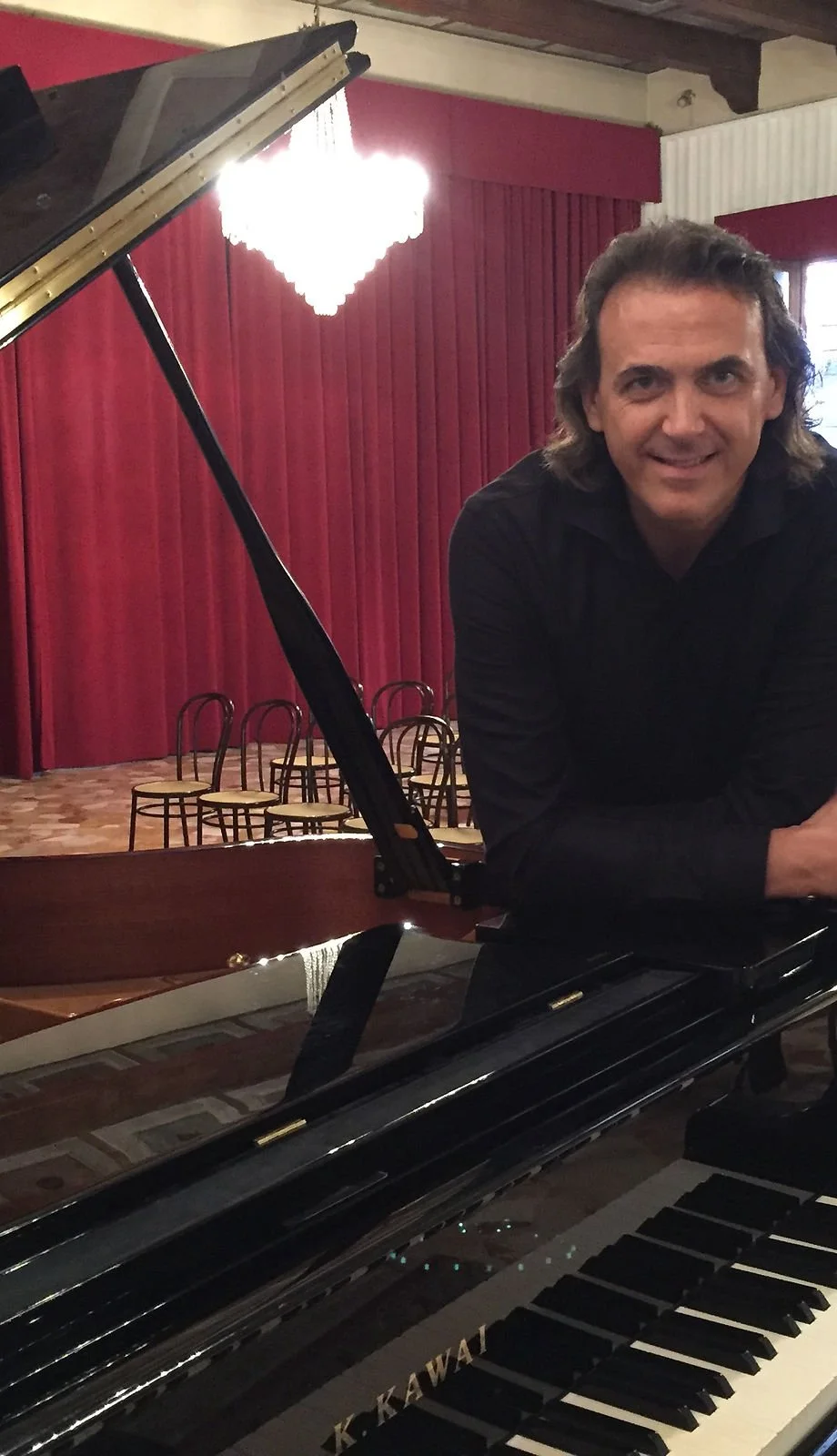 Uomo con capelli lunghi sorridente accanto a un pianoforte a coda nero in un teatro con pareti rosse e sedie vuote sul palco.