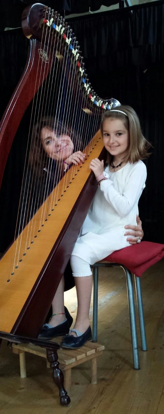 Una ragazza seduta su una sedia con un harpsichord, sorridente e con una mano sulla tastiera, con una bambina dietro di lei che si affaccia tra le corde dell'instrumento, entrambi sorridenti.