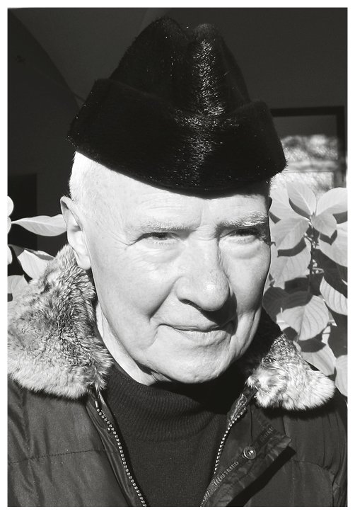 Un uomo anziano con cappello nero, abbigliamento invernale e sfondo di foglie. L'immagine è in bianco e nero.