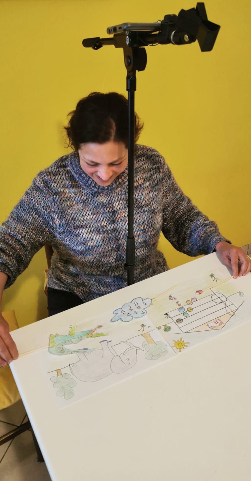 Una donna sorridente si rende conto di essere ripresa con una telecamera posta sopra la testa, mentre guarda un disegno colorato su un tavolo.